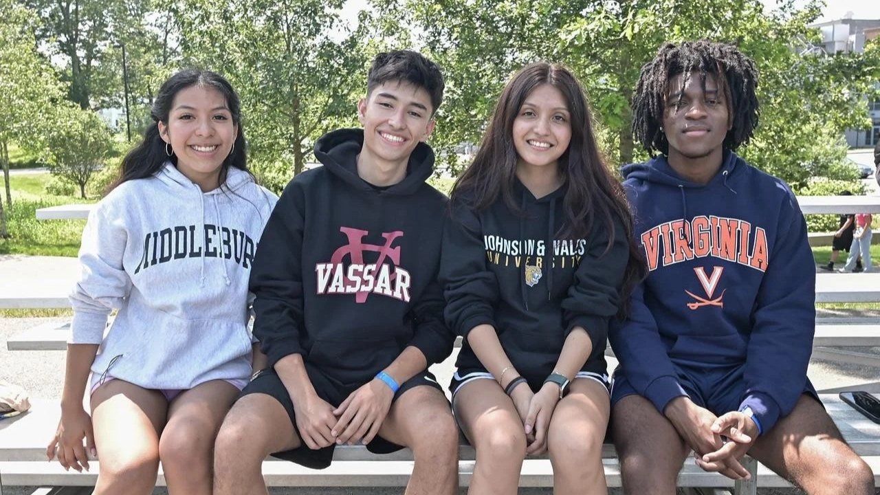 Group of four, diverse young people wearing their college sweatshirts. They are sitting on a white bench outdoors in a college campus setting.