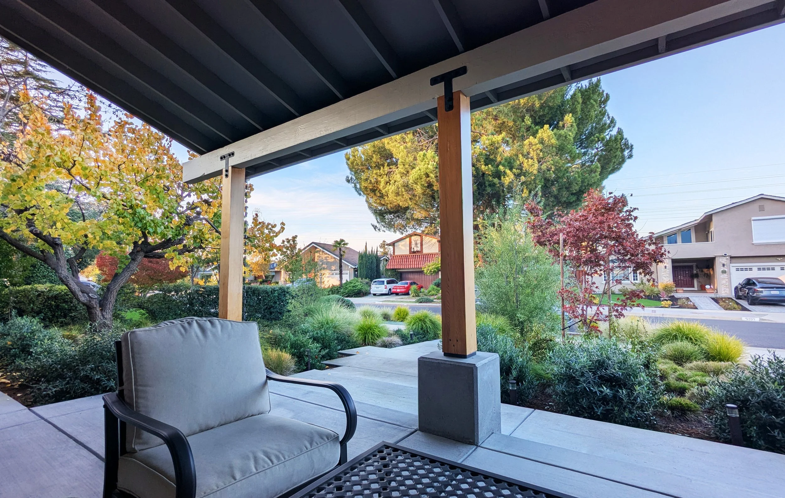 View of a residential front yard garden, from the front porch area; there are shrubs and trees in the background and a neighborhood street.
