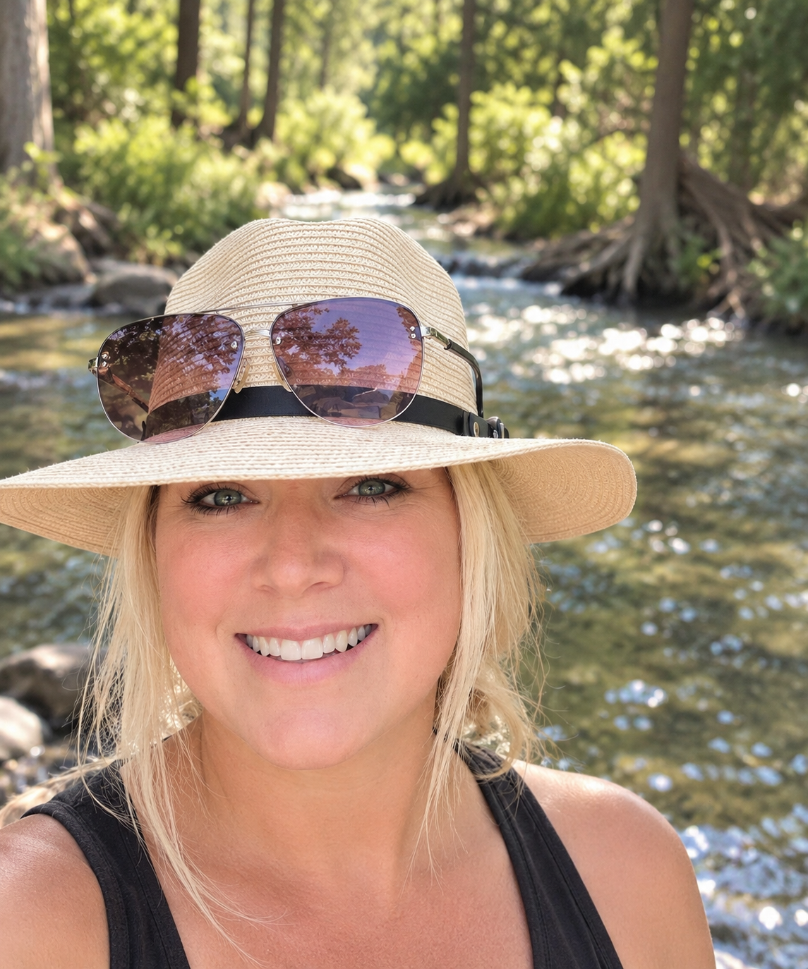 A woman wearing a wide-brimmed straw hat with sunglasses on it, smiling at the camera, outdoors by a creek, surrounded by trees and lush greenery.