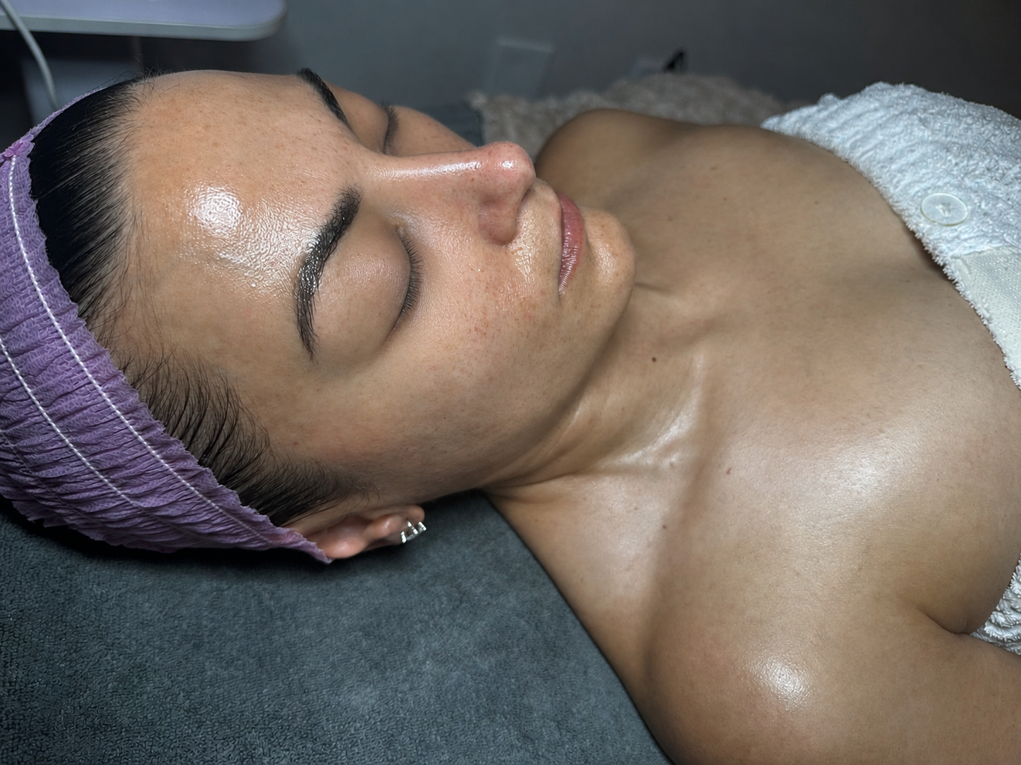 A woman lying on her back with her eyes closed, wearing a purple hair cap, in a spa or skincare setting.