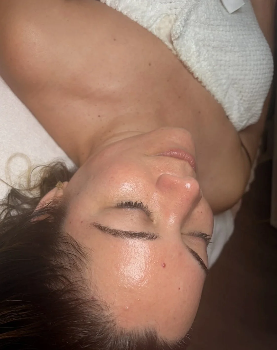A woman lying on a bed with her eyes closed, covered partially with a white towel or cloth, appears to be relaxing or receiving a spa treatment.
