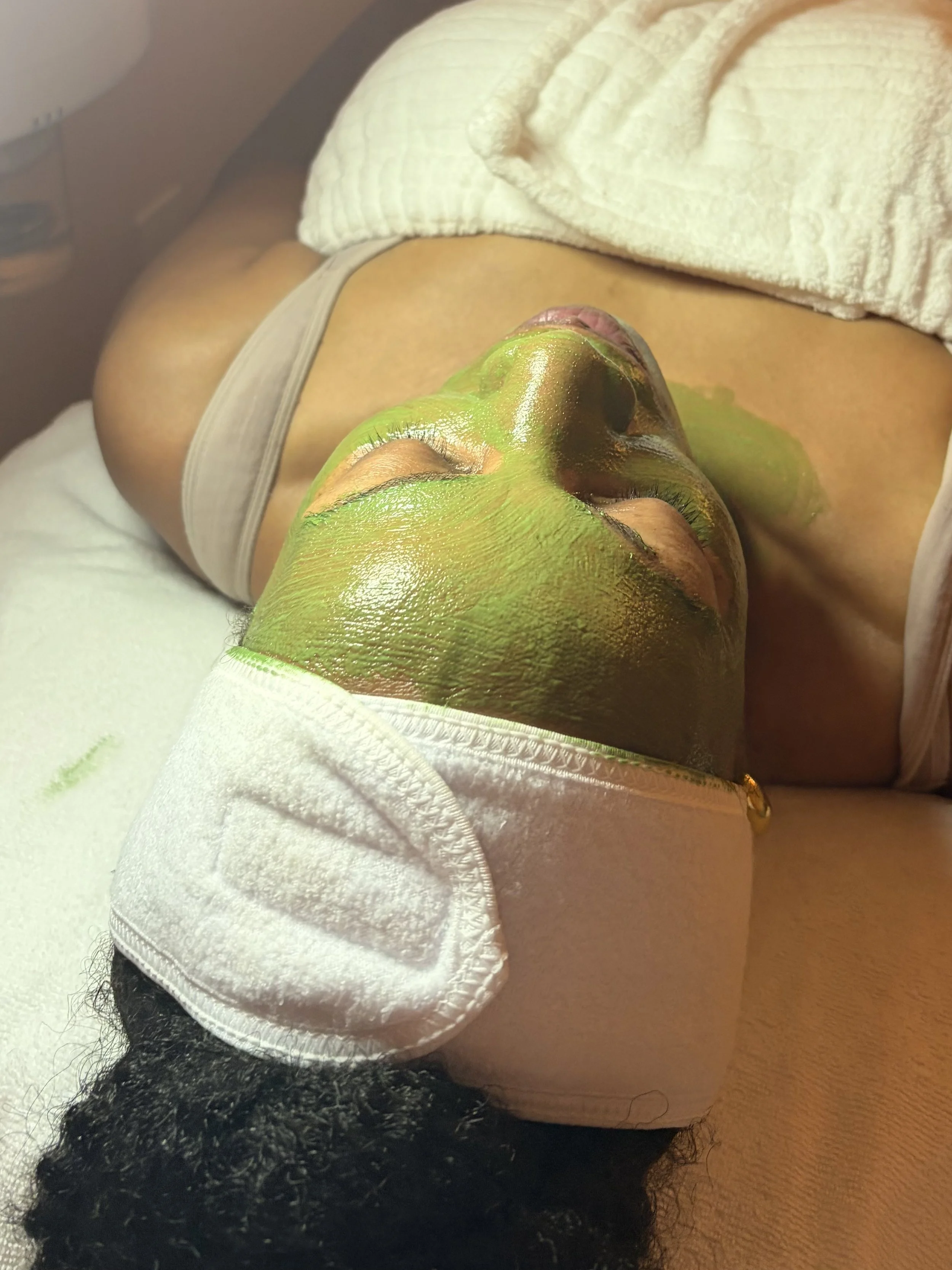 A person lying on their back with green face paint, curly black hair, and closed eyes, in a spa or treatment setting with a towel covering their chest.
