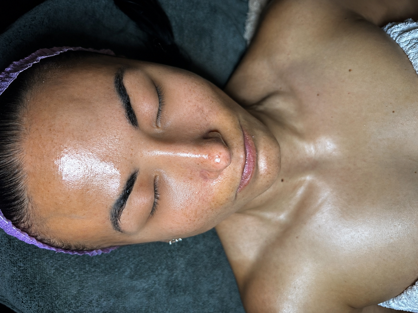 A woman with dark hair, light brown skin, closed eyes, and a relaxed expression, lying down with a towel on her head, after a facial treatment.
