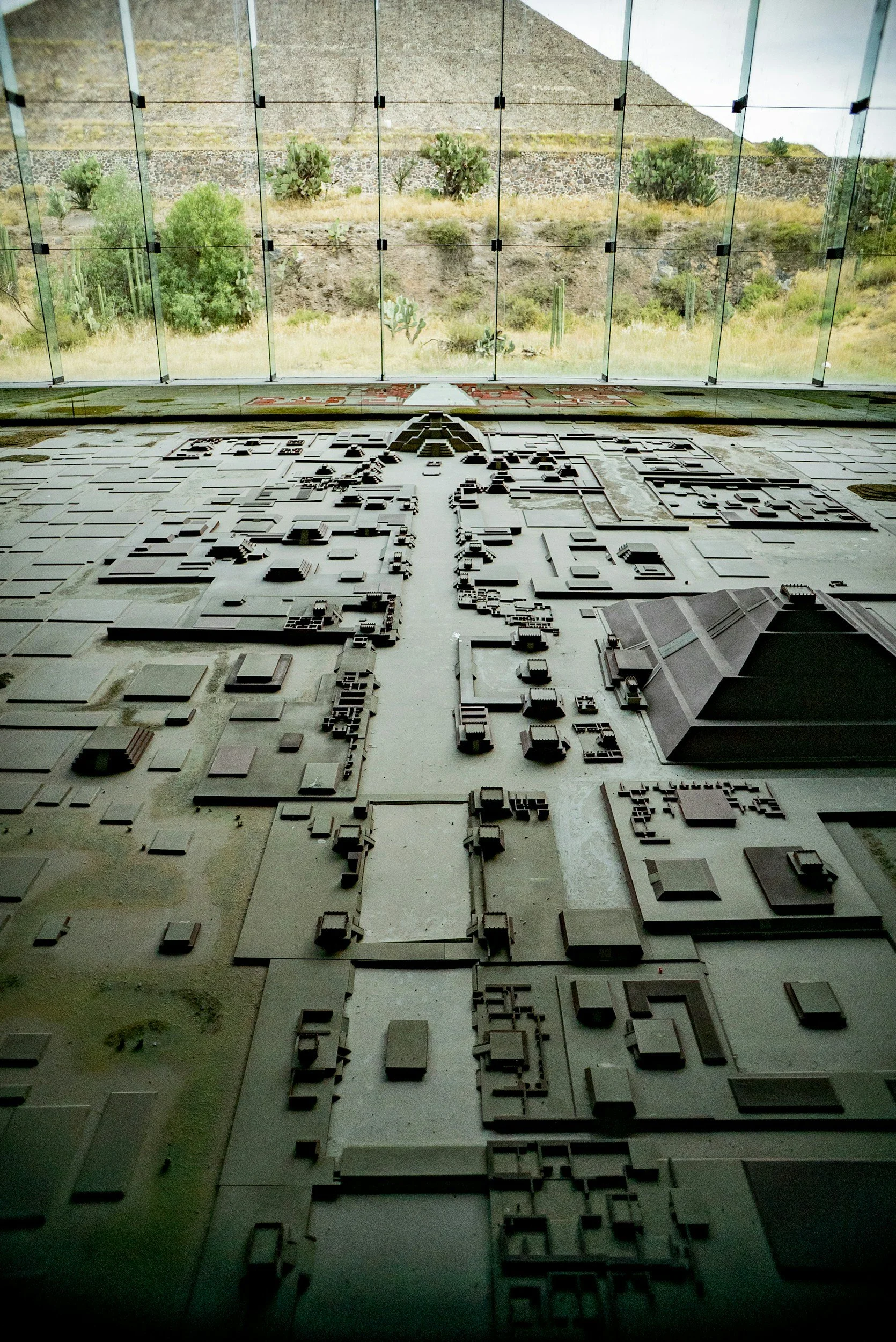 Scale model of an ancient city with buildings, streets, and a pyramid-like structure, viewed from above, with large glass windows showing a desert landscape with cacti outside.