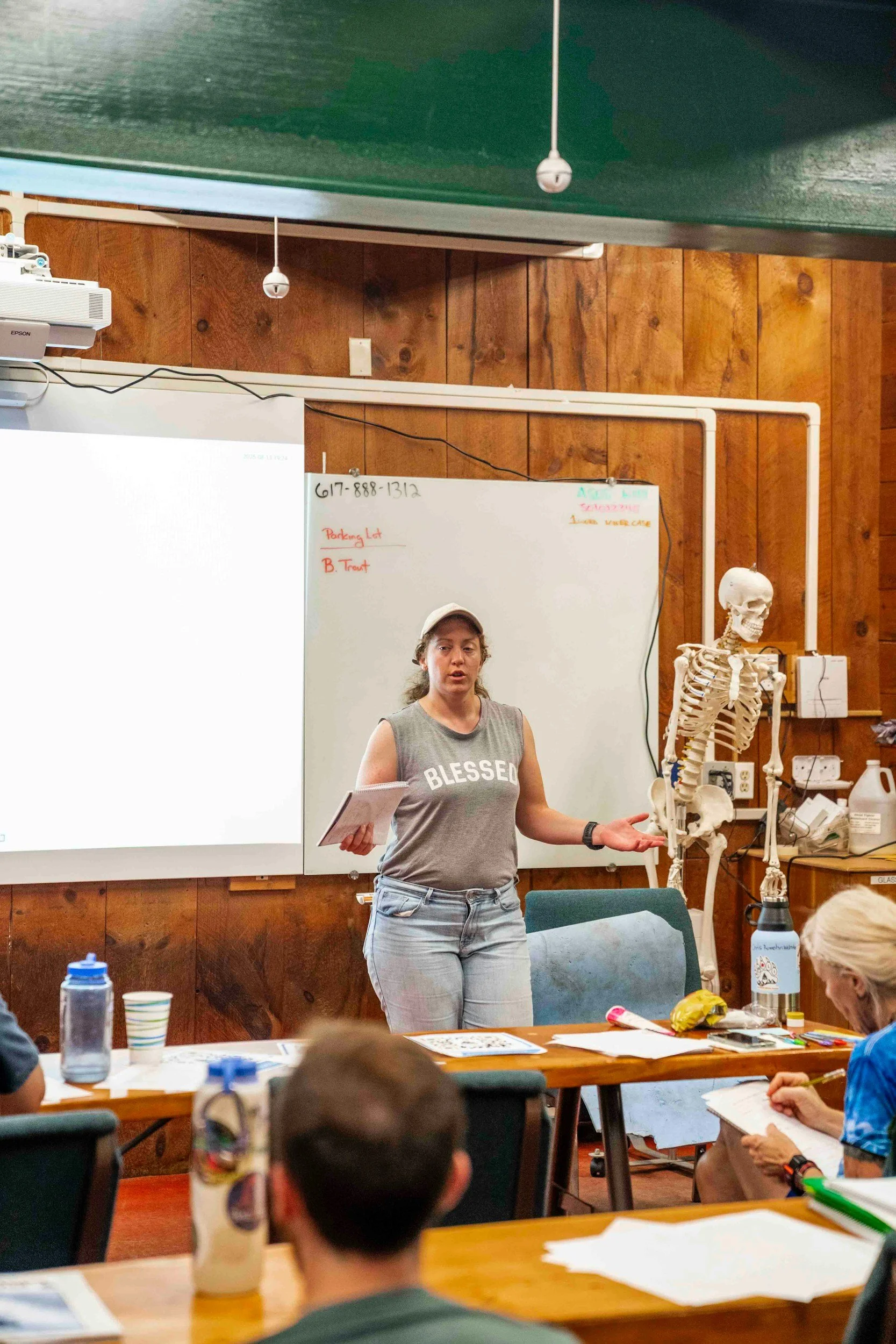 A woman standing in front of a classroom or meeting room, gesturing while speaking, with a skeleton model beside her, and a whiteboard, projector, and audience seated at tables with notebooks and drinks.