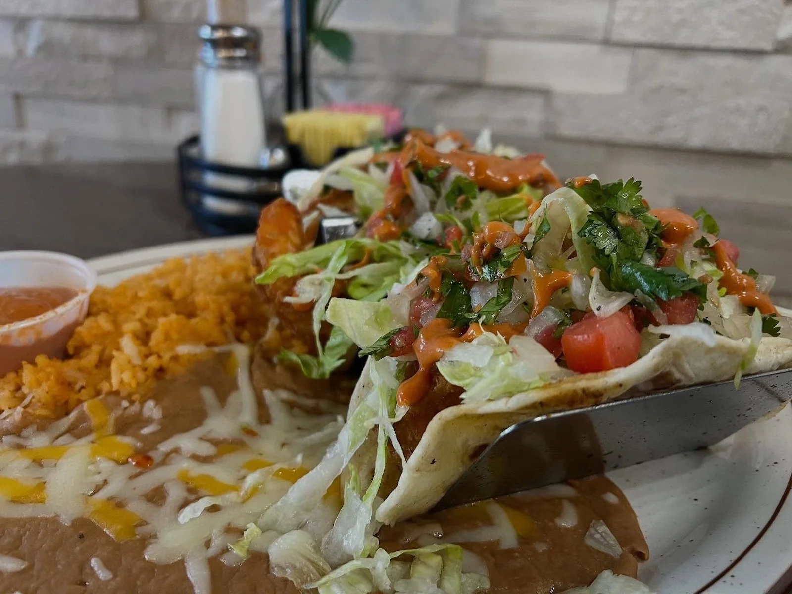 A plate of Mexican food featuring a taco filled with lettuce, tomatoes, cilantro, and sauce, served with rice, beans, cheese, and a small side of salsa.