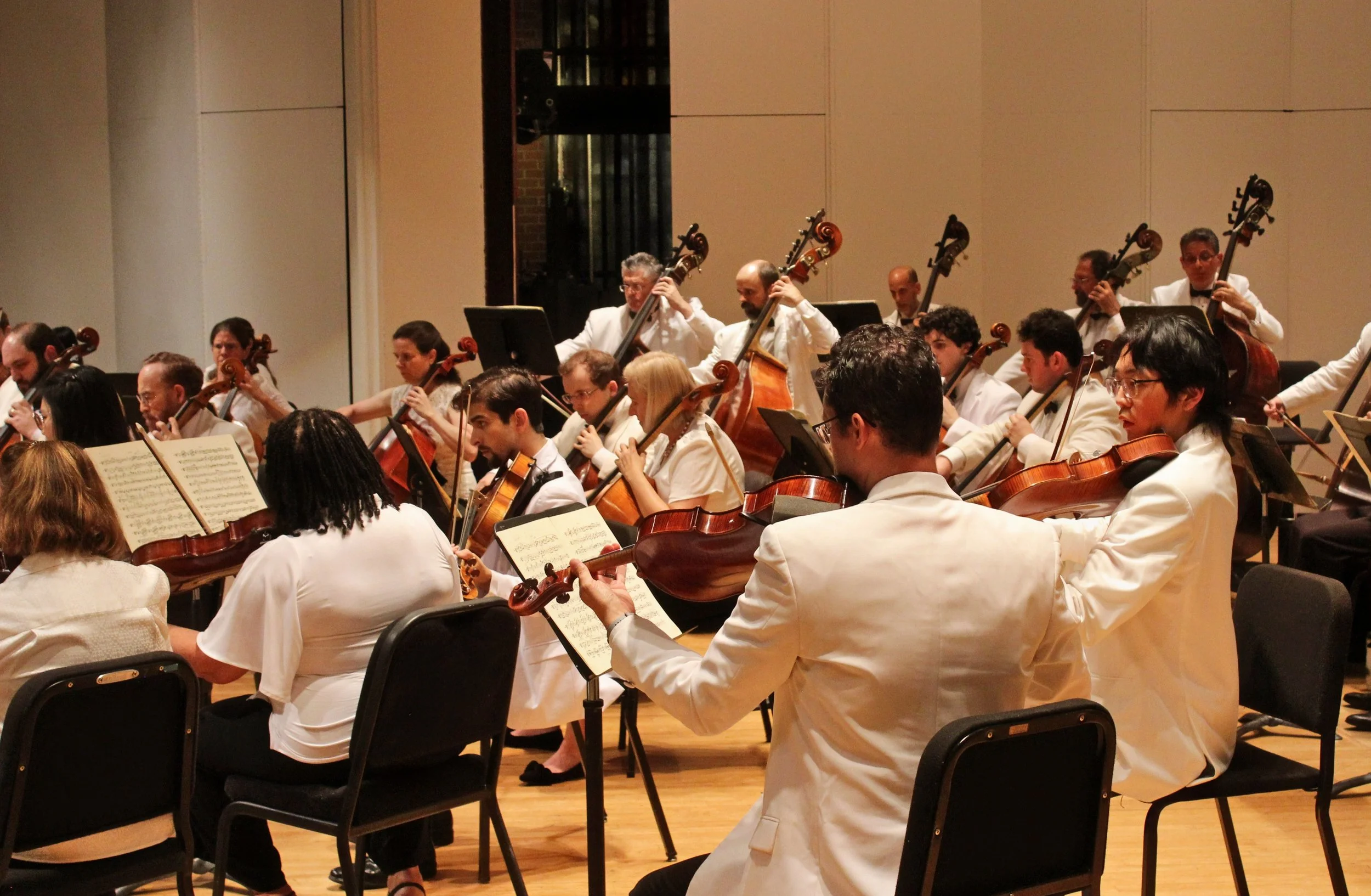 The Eastern Festival Orchestra performing on stage, with musicians playing string instruments like violins and cellos. The performers are dressed in formal white attire, and music sheets are in front of them.