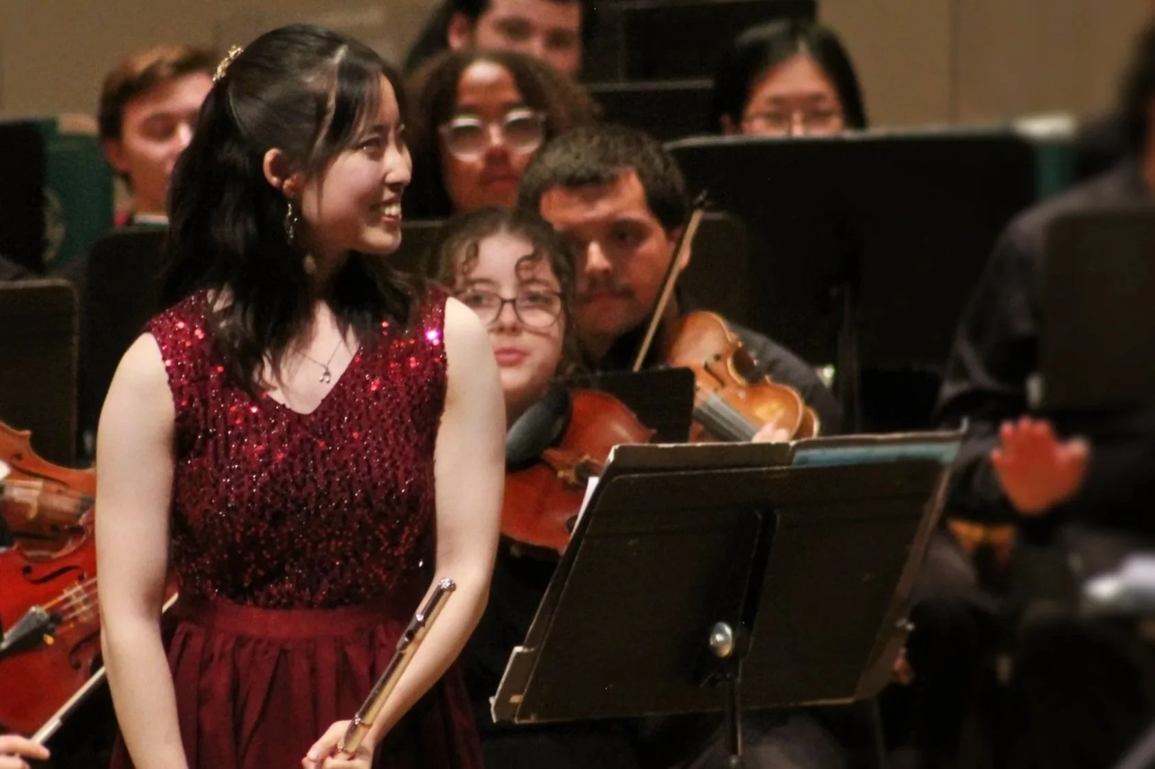 Young woman in a red, sparkly dress holding a flute, smiling, surrounded by musicians, some with violins, during an orchestra performance.