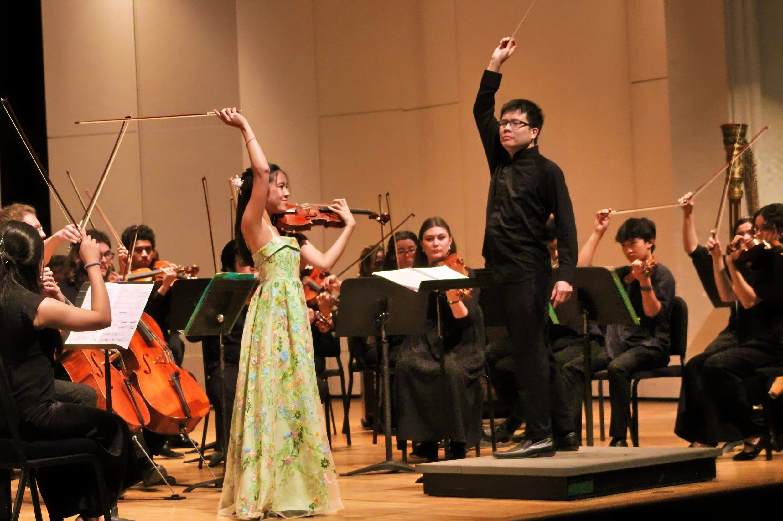 An orchestra performing on stage with a woman in a floral dress playing the violin and Ho-Yin Kwok conducting.