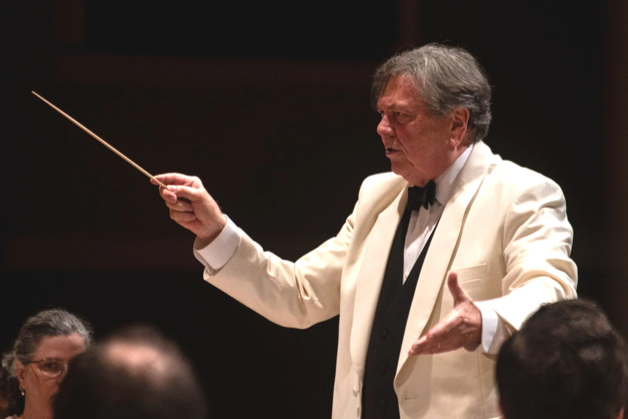 Gerard Schwarz, dressed in a white tuxedo and black bow tie, conducting an orchestra with a baton.