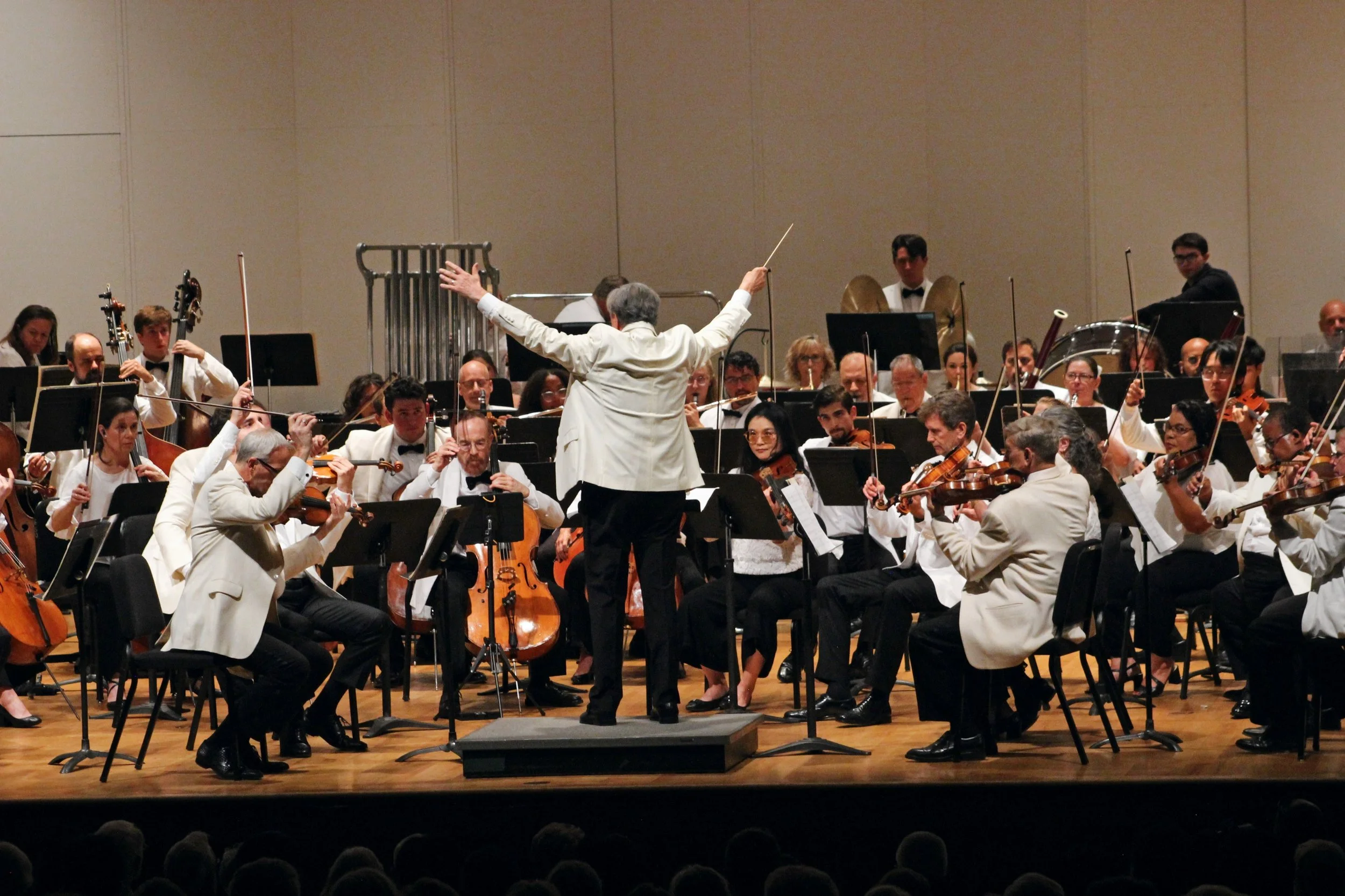 Orchestra performing on stage, Gerard Schwarz conducting and leading, musicians playing string and percussion instruments, audience in foreground.
