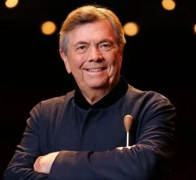 Gerard Schwarz, smiling and wearing a dark shirt, standing in front of a dark background with blurred lights.