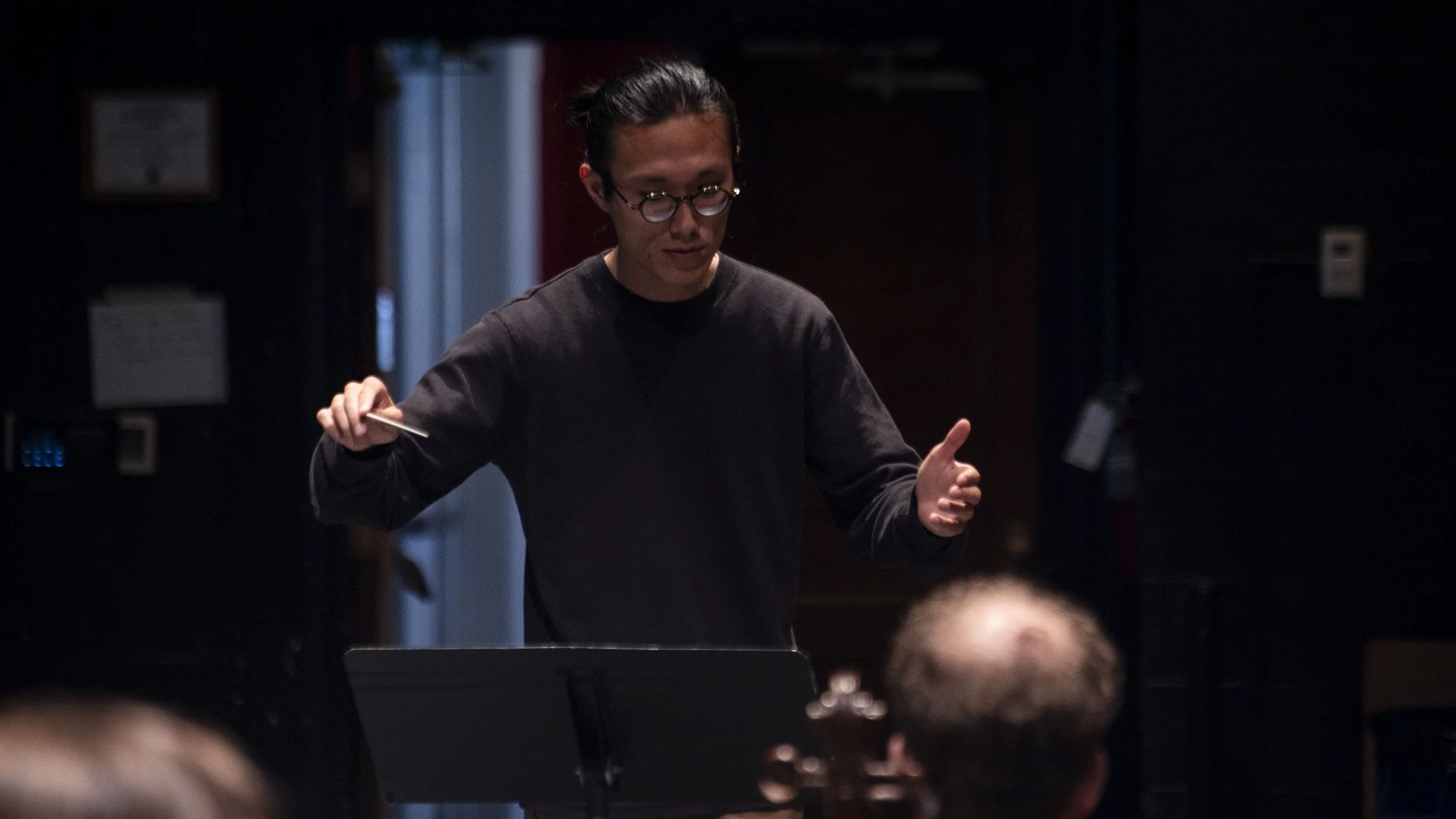 A person with glasses and dark hair styled in a bun, wearing a dark long-sleeve shirt, is conducting or leading an activity indoors, with a dark background and a music stand in front of them.