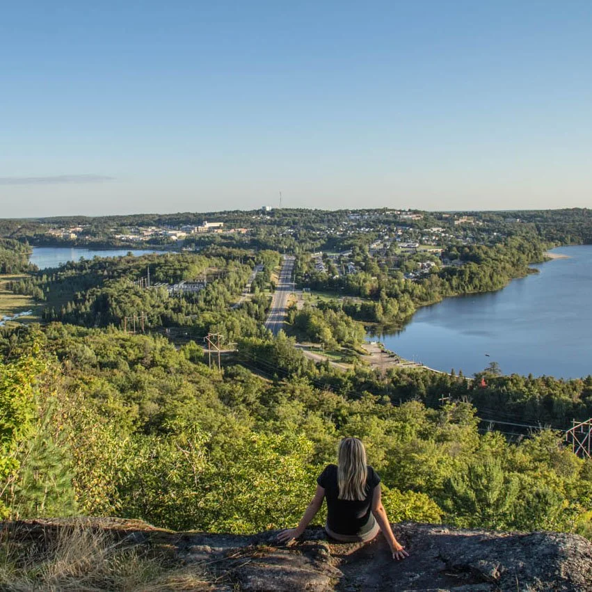 A woman sitting on a rock at a high vantage point overlooking a scenic view of lakes, greenery, and a distant town under a clear blue sky.