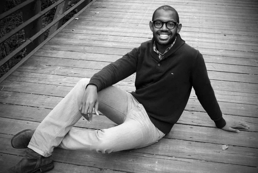 A man with glasses, beard, and short hair sitting on a wooden bridge, smiling at the camera, wearing a dark sweater, light pants, and shoes.