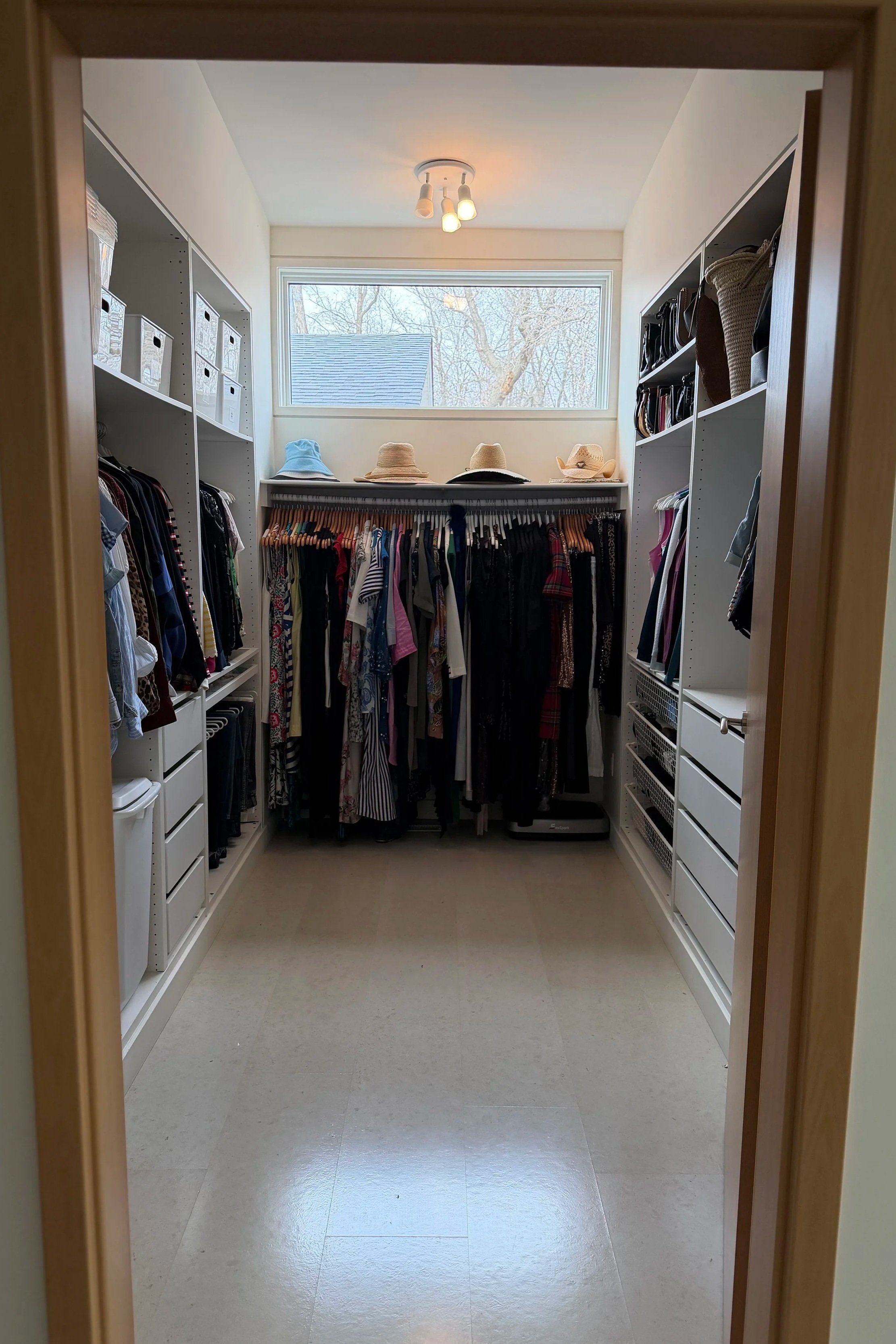 Walk-in closet with white shelving, hanging clothes, storage baskets, hats, and a large window.