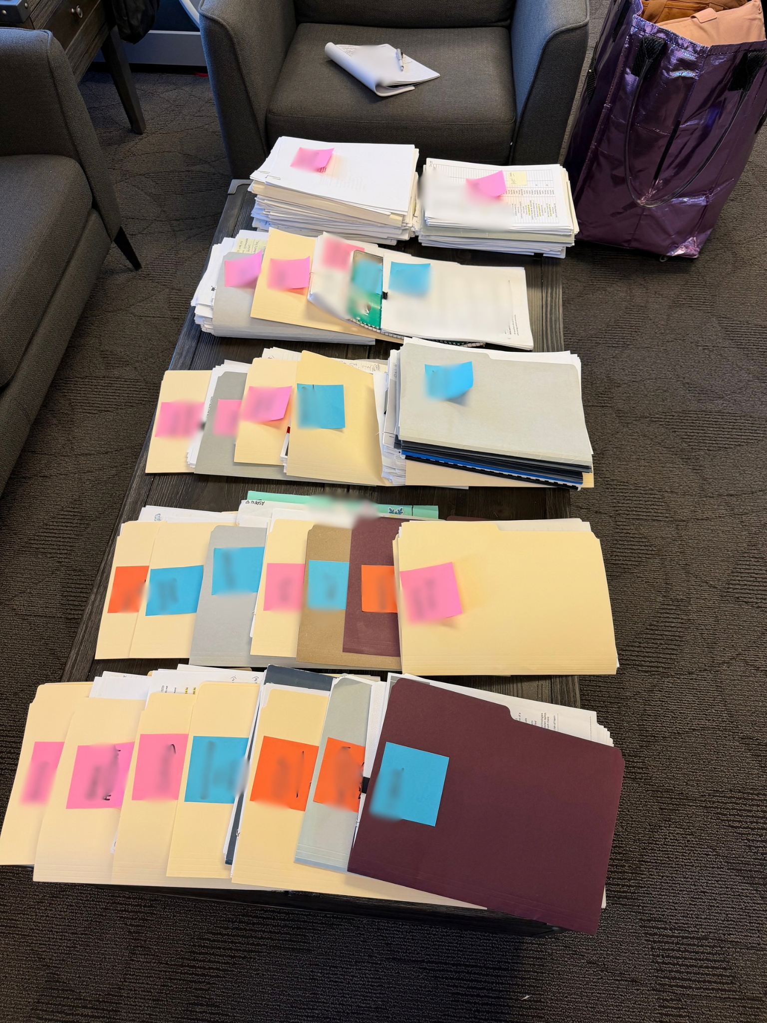 Stacks of organized paperwork and file folders with colorful sticky notes on a table beside a purple tote bag