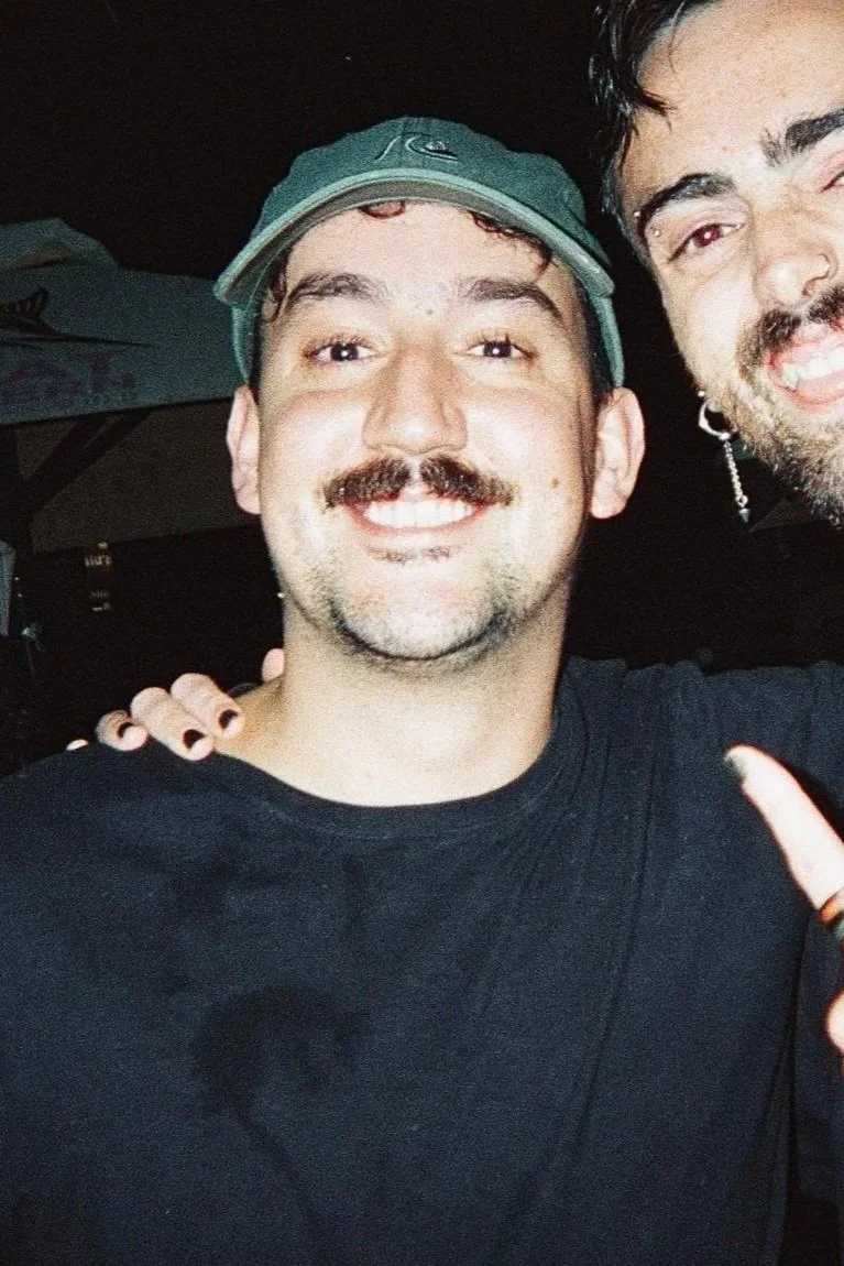 Two men smiling for a selfie indoors, one wearing a baseball cap and the other with dark hair and a beard, with their faces close together.