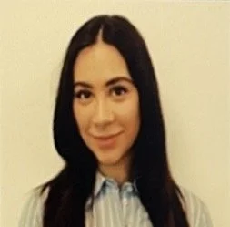 A woman with long dark hair smiling at the camera, wearing a light-colored collared shirt against a plain background.