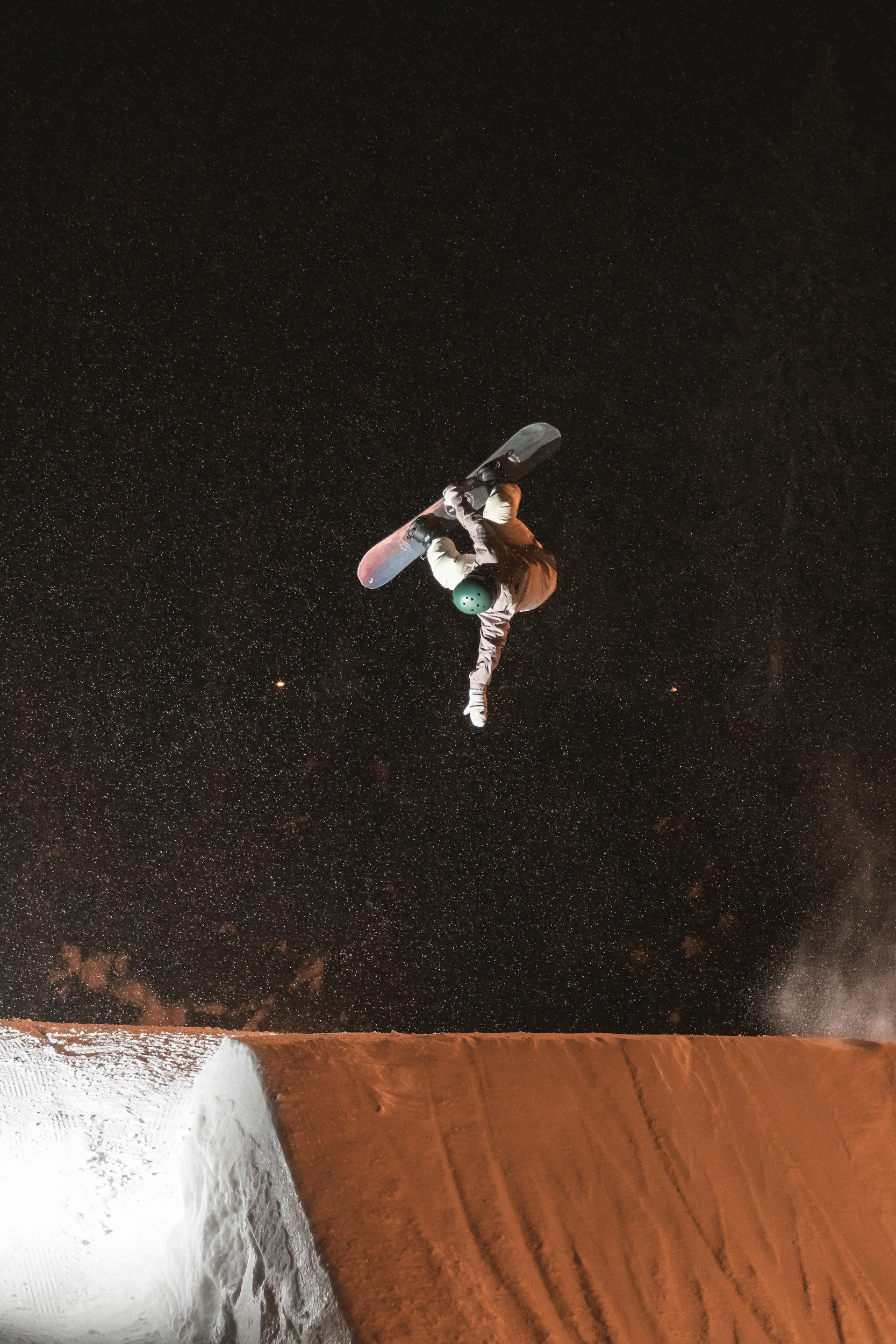 A skateboarder performing a trick in mid-air at night, with a starry sky in the background and snow-covered ground and a snowbank at the bottom.