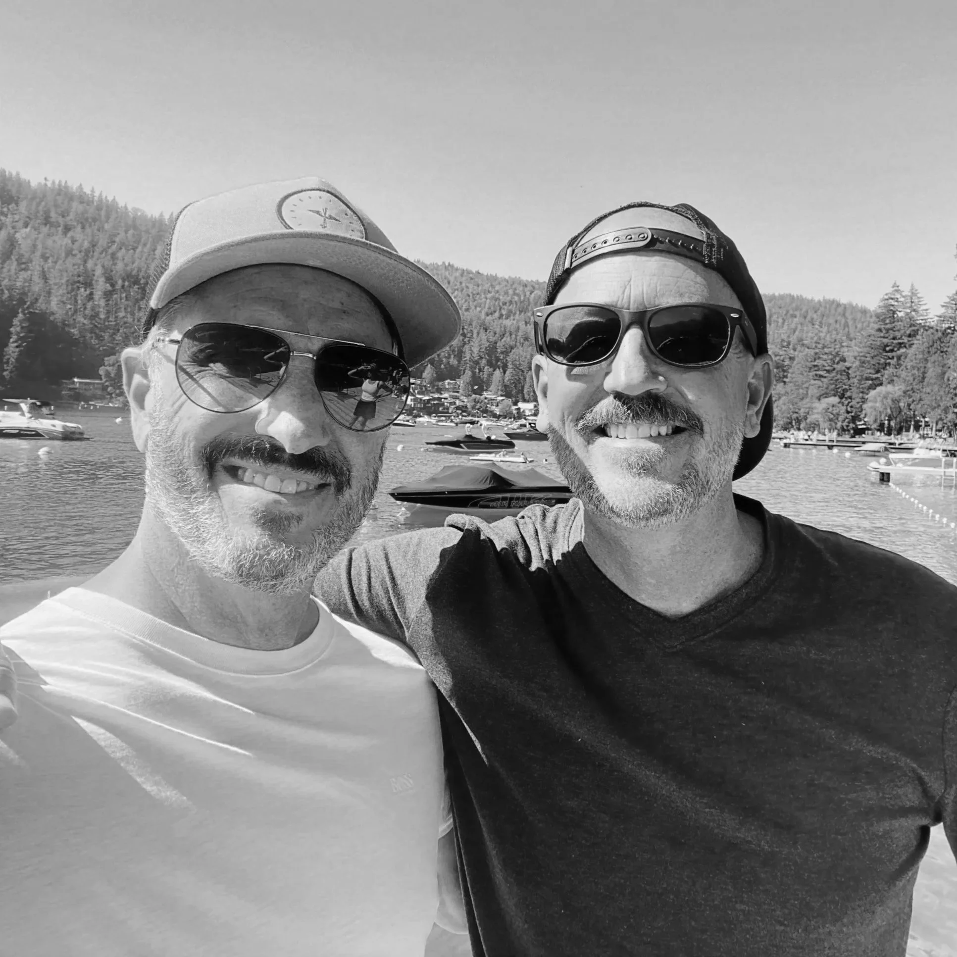 Black and white photo of two men smiling, wearing sunglasses and caps, with a lake, boats, and forested mountains in the background.
