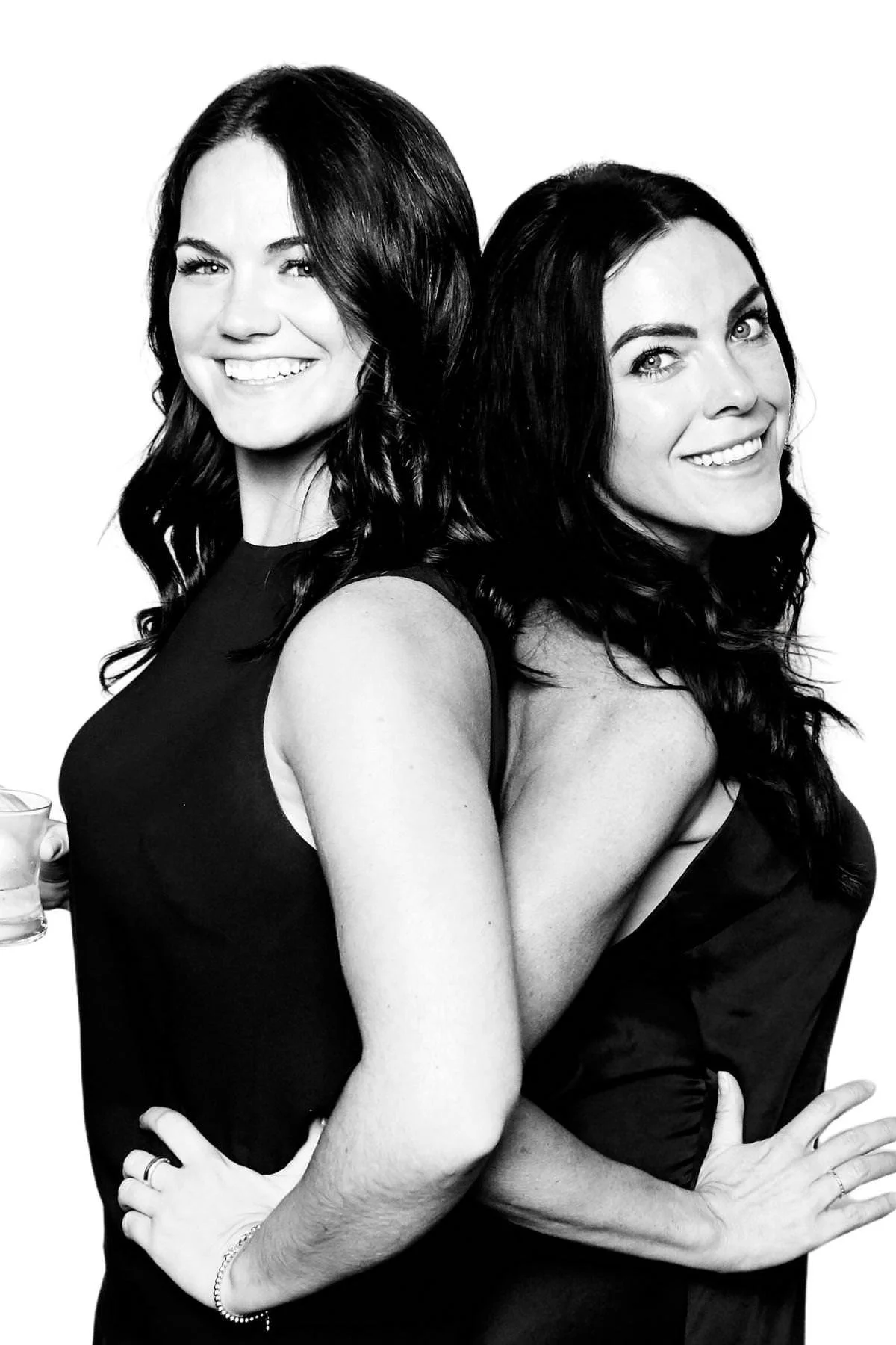 Two smiling women in black dresses posing back to back on a white backdrop