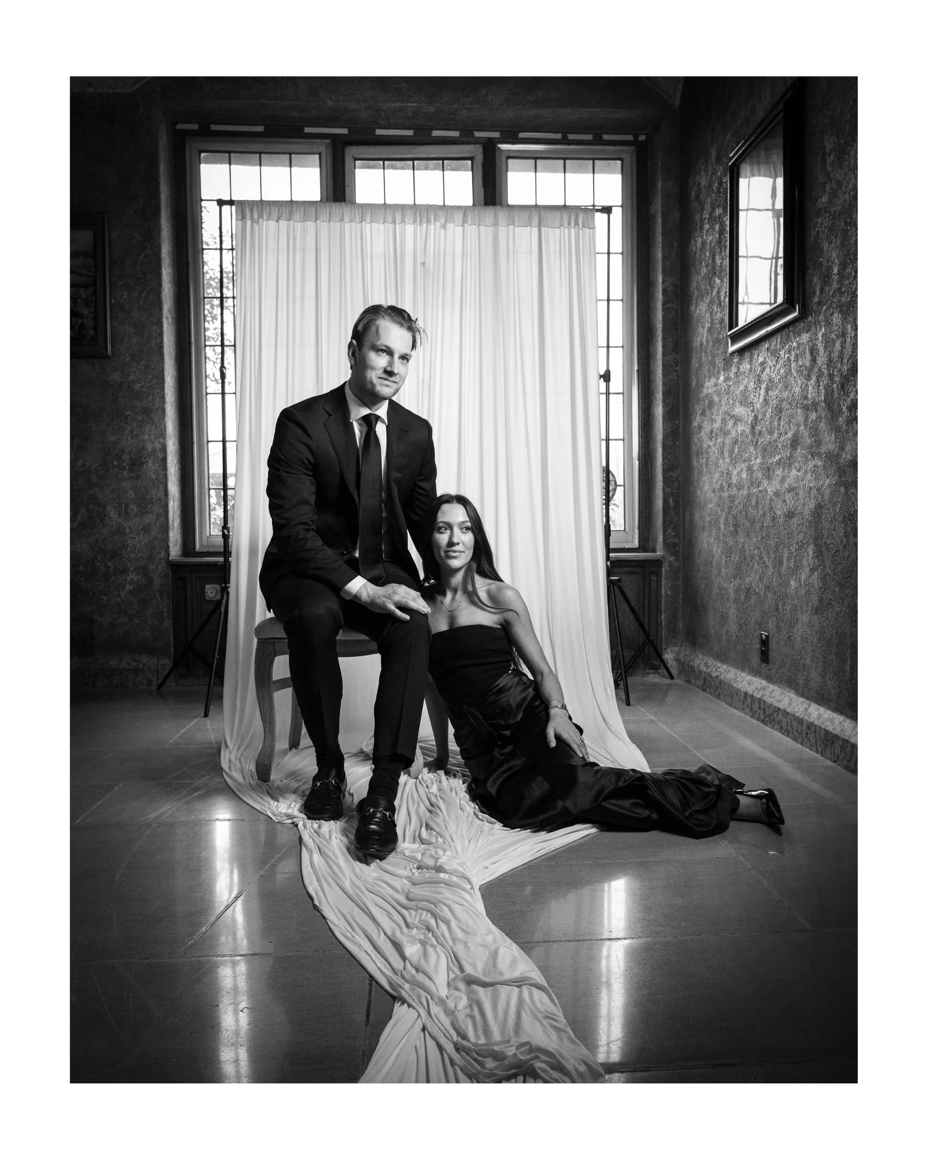 Black and white portrait of a man seated on a chair with a woman in a black gown sitting at his feet on a white drape
