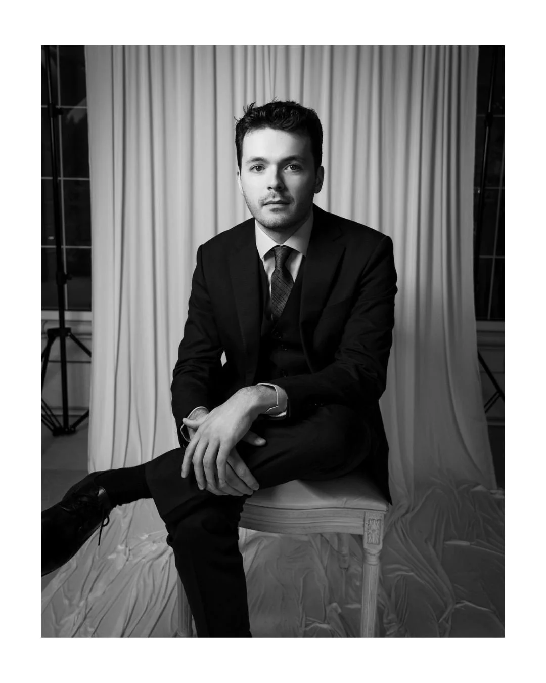 Black and white portrait of a young man in a suit seated in front of a draped backdrop