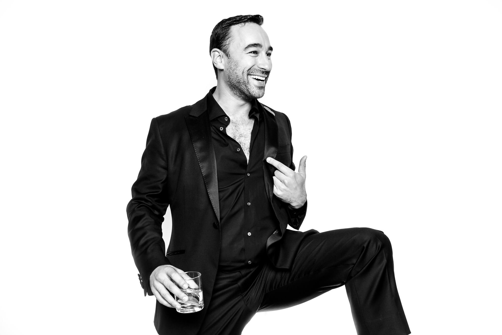 Laughing man in a black tuxedo holding a whisky glass, pointing to himself with one knee raised
