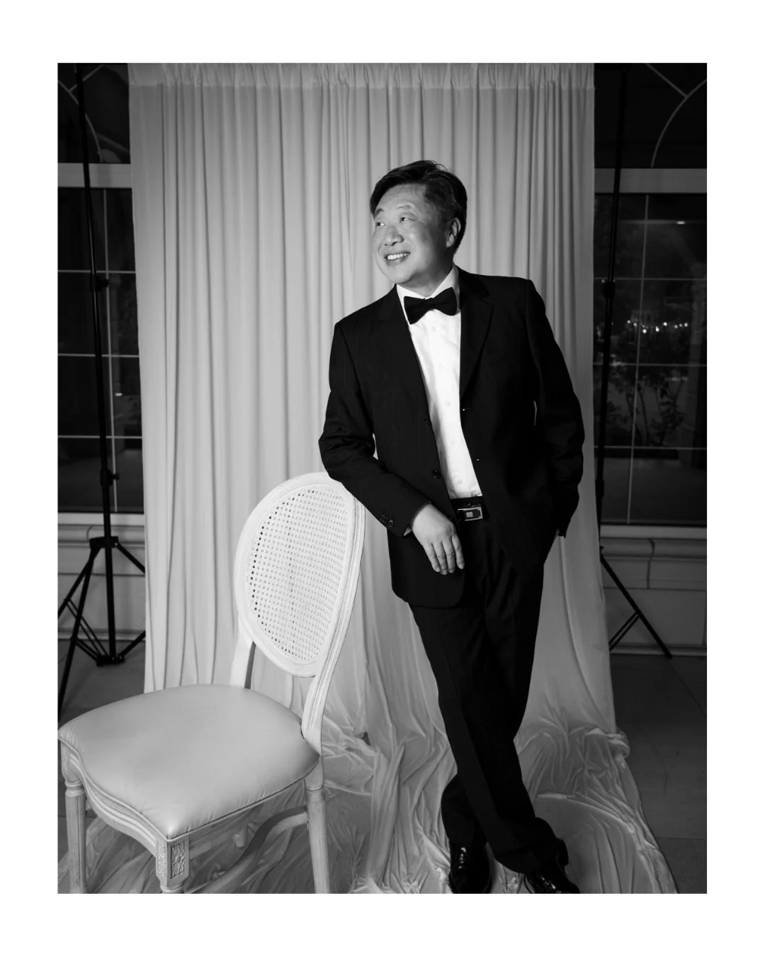 Black and white portrait of a smiling guest in a tuxedo and bow tie leaning on a cane-back chair at Archway Manor
