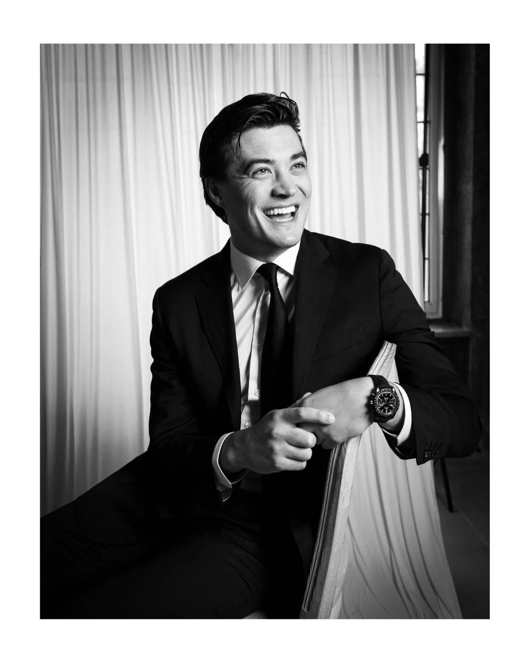 A smiling man in a suit sitting on a chair, posing for a portrait, with a light studio backdrop in the background.