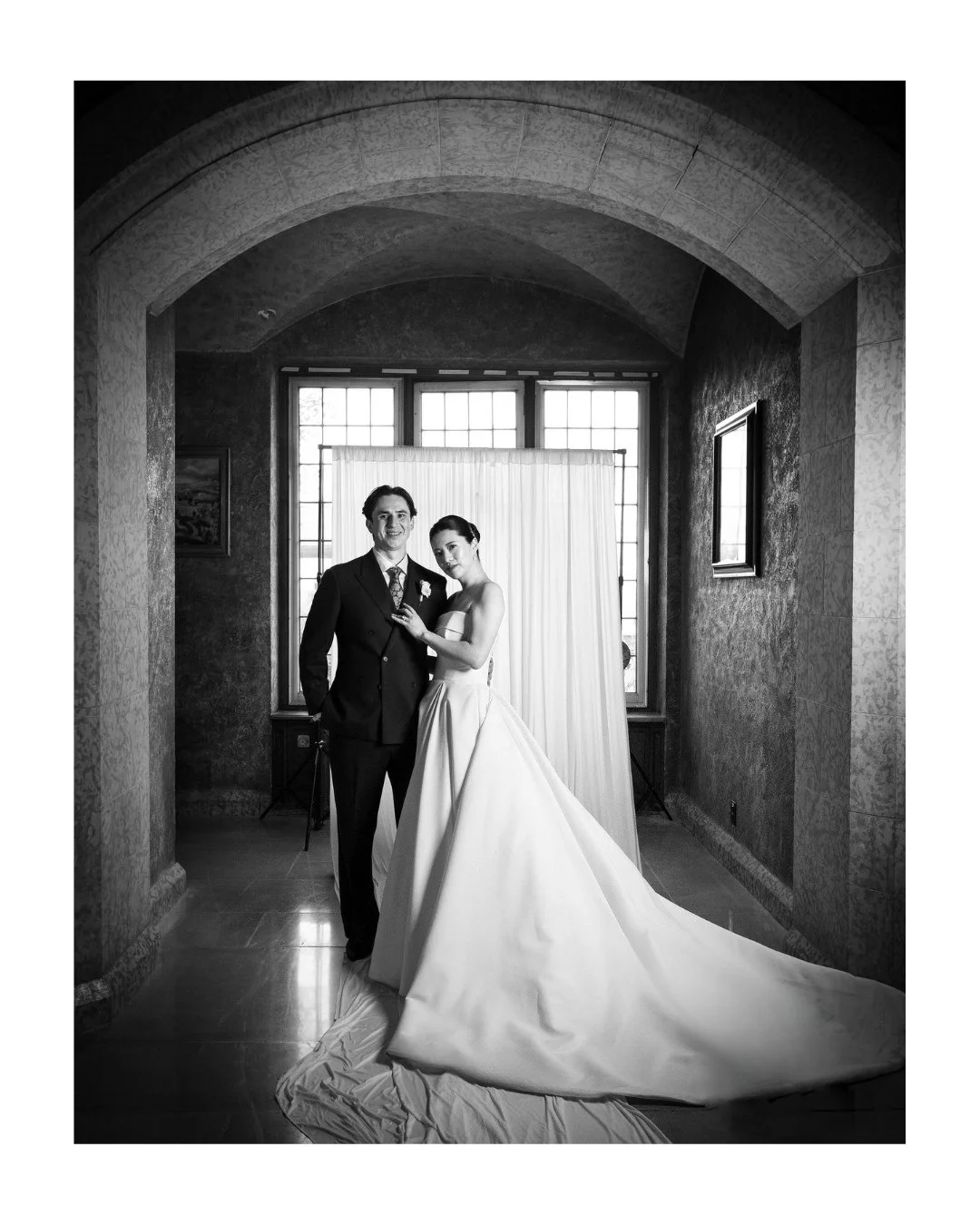 Black and white portrait of a bride and groom in a stone archway, framed by a draped backdrop and tall windows