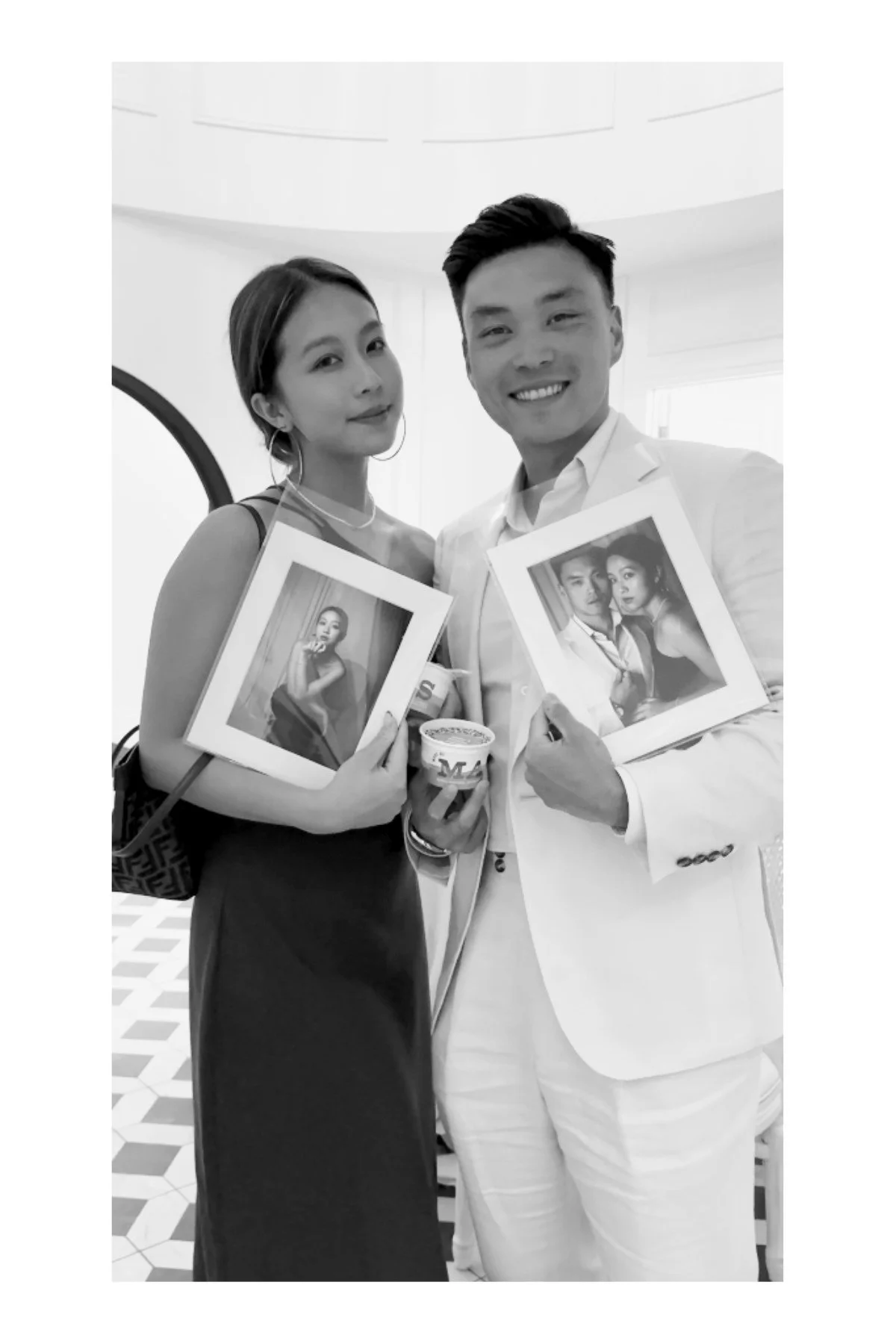 Black and white photo of a man and woman dressed in formal attire, standing indoors and smiling at the camera. The woman is holding a framed photograph of herself, and the man is holding a framed photograph of himself and her together, along with a cup.