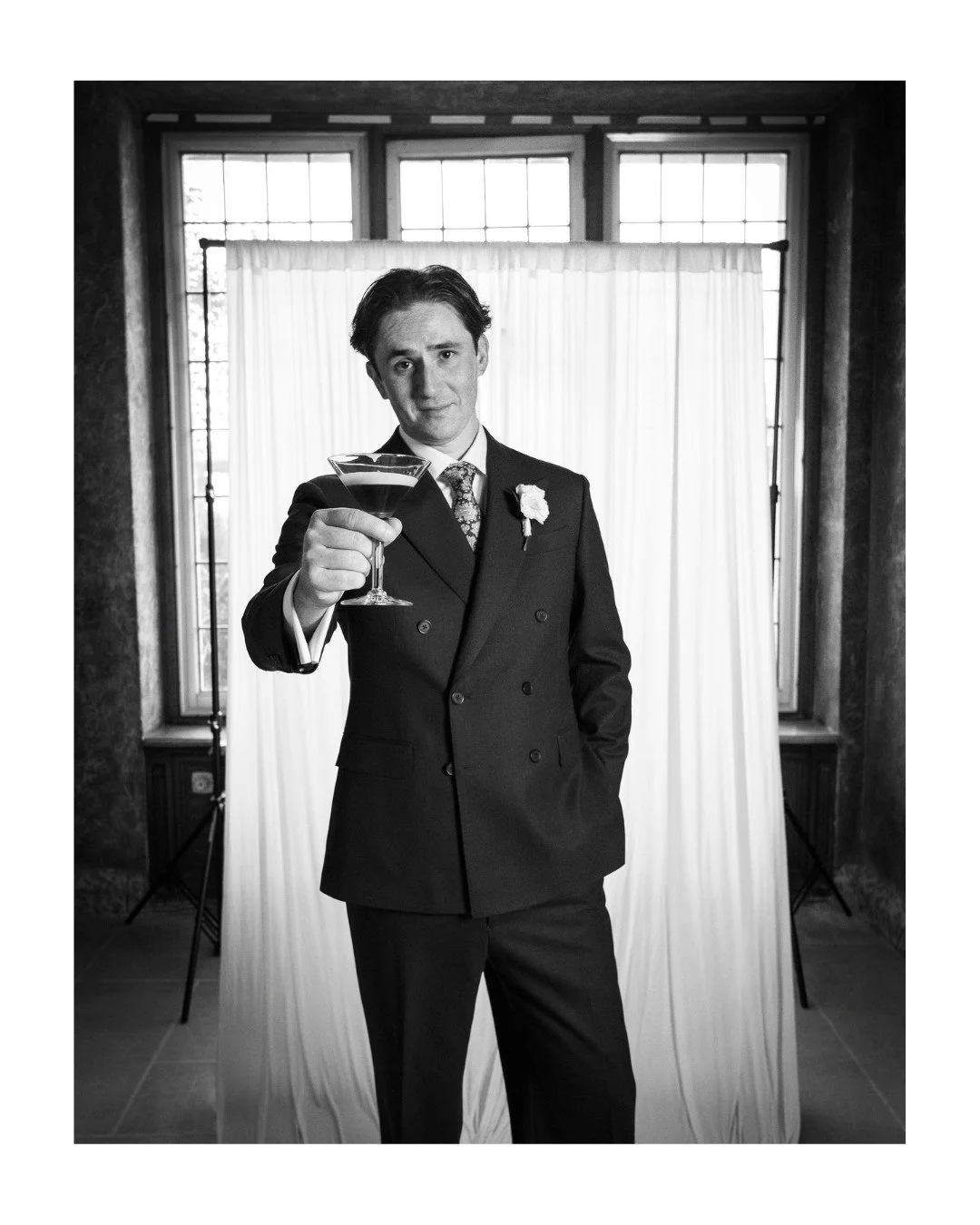 Black and white portrait of a groom holding a cocktail in front of a window and draped backdrop