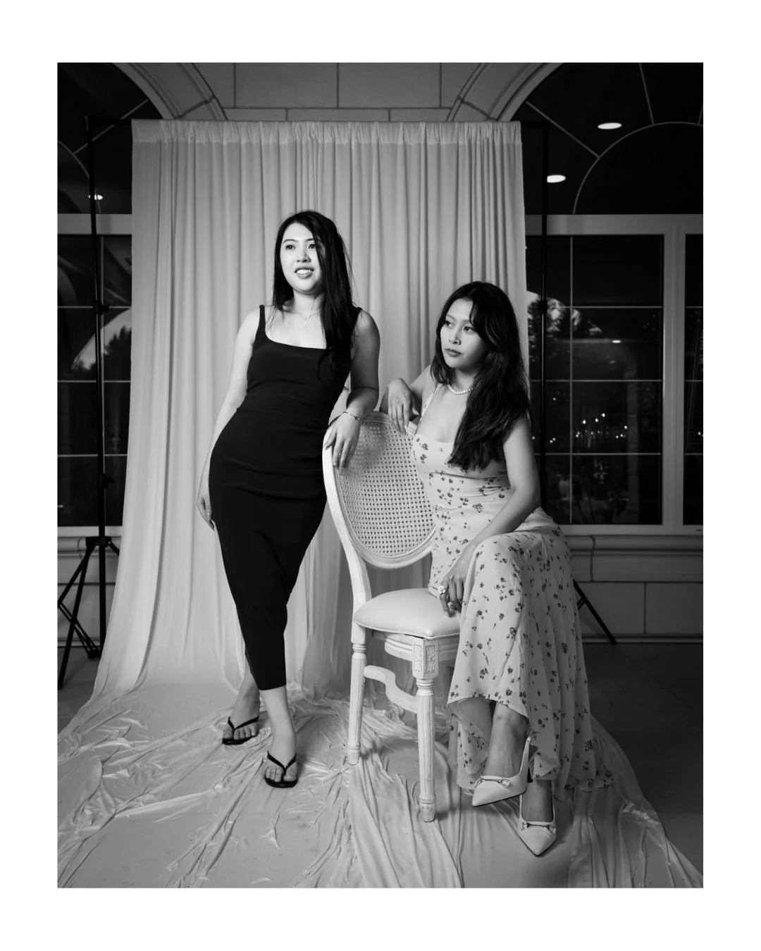 Two wedding guests posing in front of a draped backdrop, one seated in a floral dress