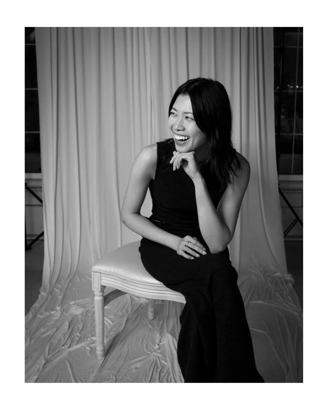 Black and white portrait of a laughing woman seated on a white chair in front of a draped backdrop