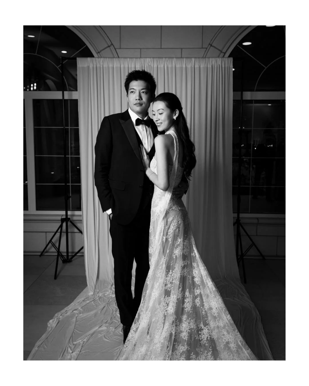 a bride and groom pose for a black and white portrait in front of a draped backdrop and stone archway.