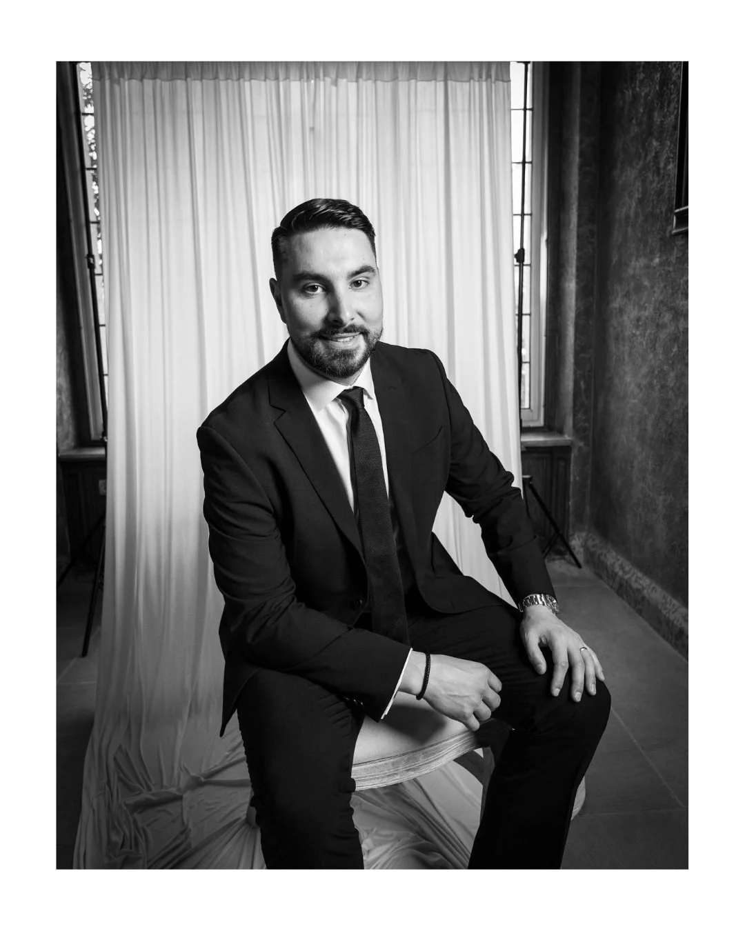 Black and white portrait of a man in a suit seated in front of a draped backdrop and window