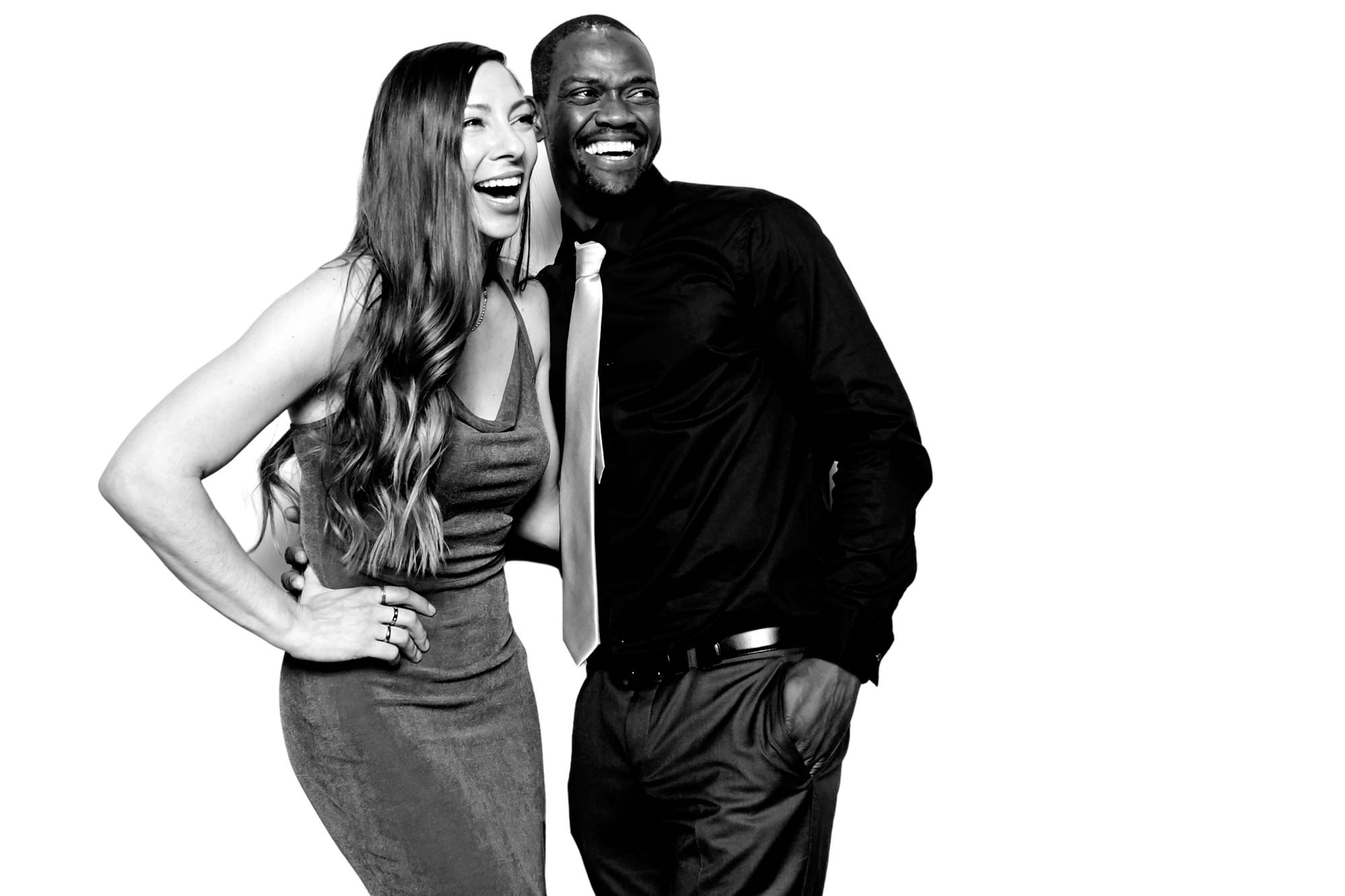 Laughing couple posing together, she in a fitted dress and he in a black shirt and tie, on a white backdrop
