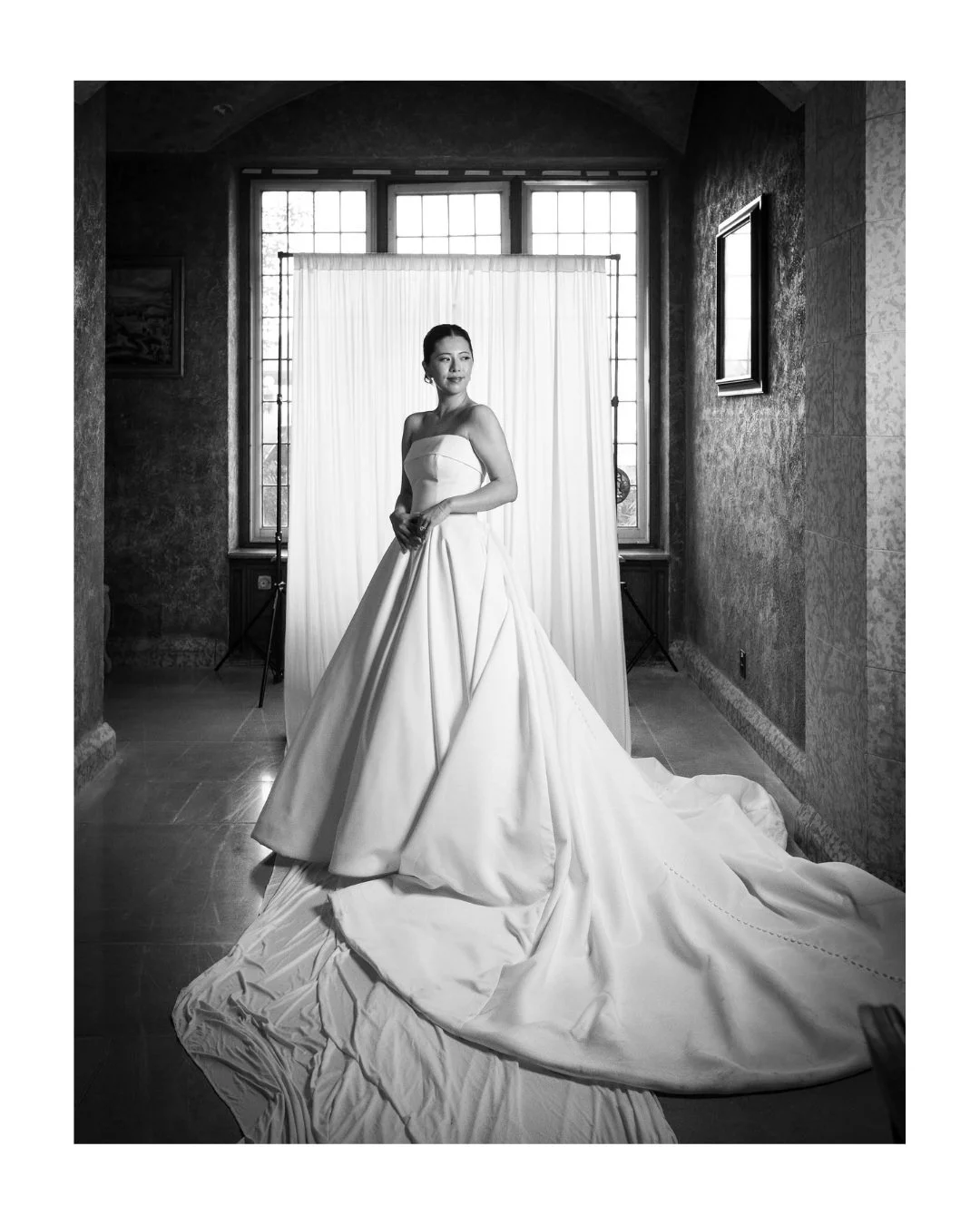 Smiling bride in her wedding gown in front of a draped backdrop and window, black and white portrait