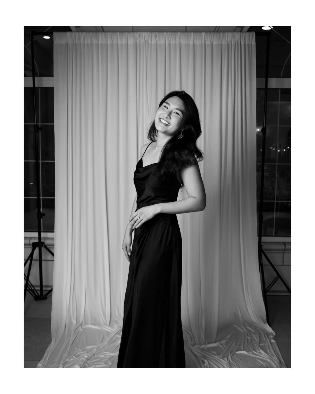Smiling woman in a long black satin dress posing in front of a light draped backdrop