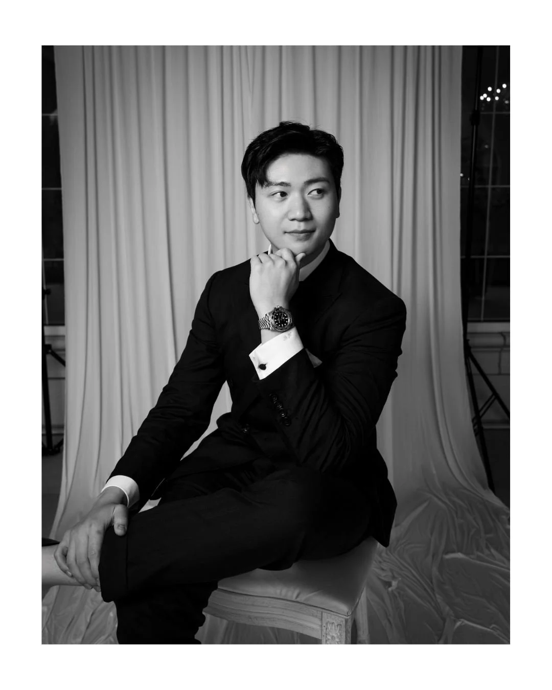 Young man in a black suit seated in front of a draped backdrop, hand on his chin