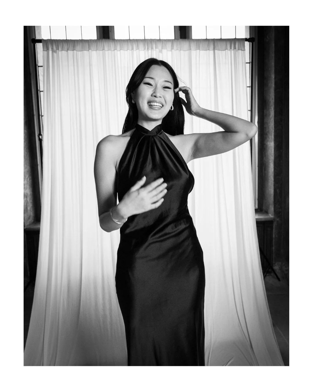 Smiling wedding guest in a sleeveless satin dress in front of a light draped backdrop