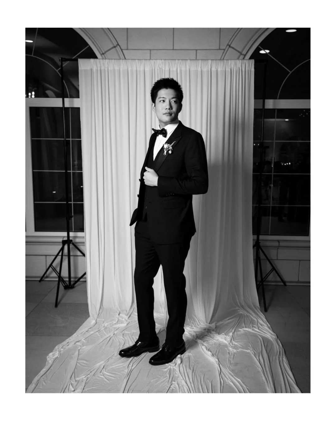 a groom poses for a black and white portrait in front of a draped backdrop and stone arches.