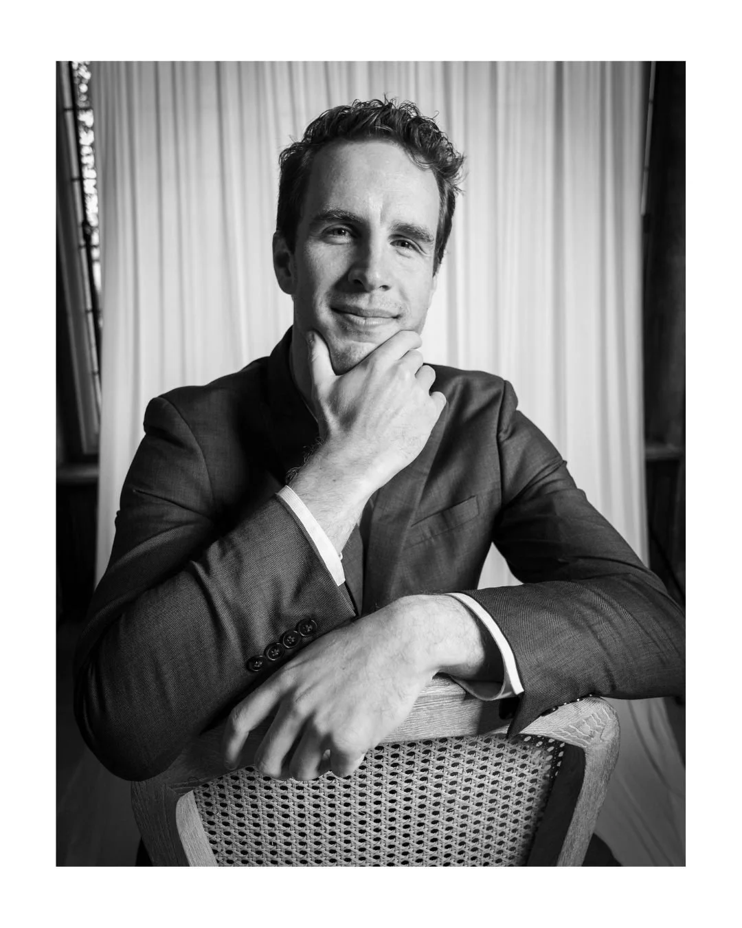 Black and white portrait of a man in a suit seated on a woven chair, chin resting on his hand