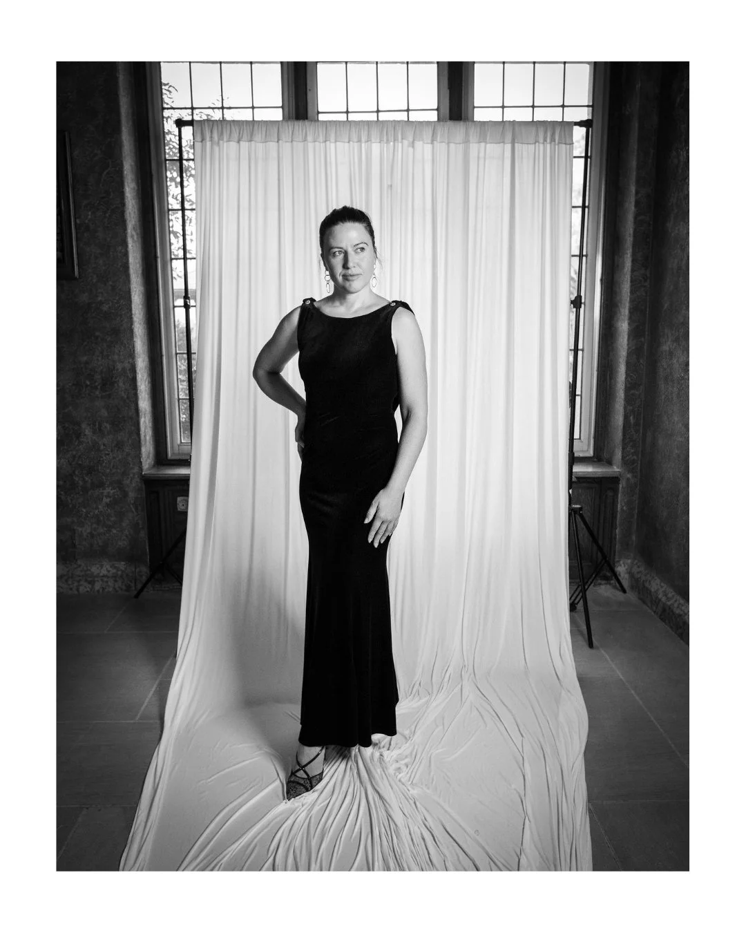 Wedding guest in a long black dress standing in front of a white draped backdrop and tall windows
