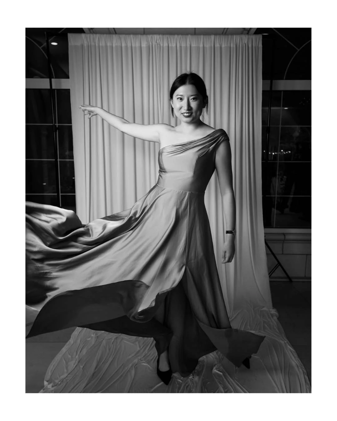 Black and white portrait of a woman in a satin gown in front of a draped backdrop and tall windows