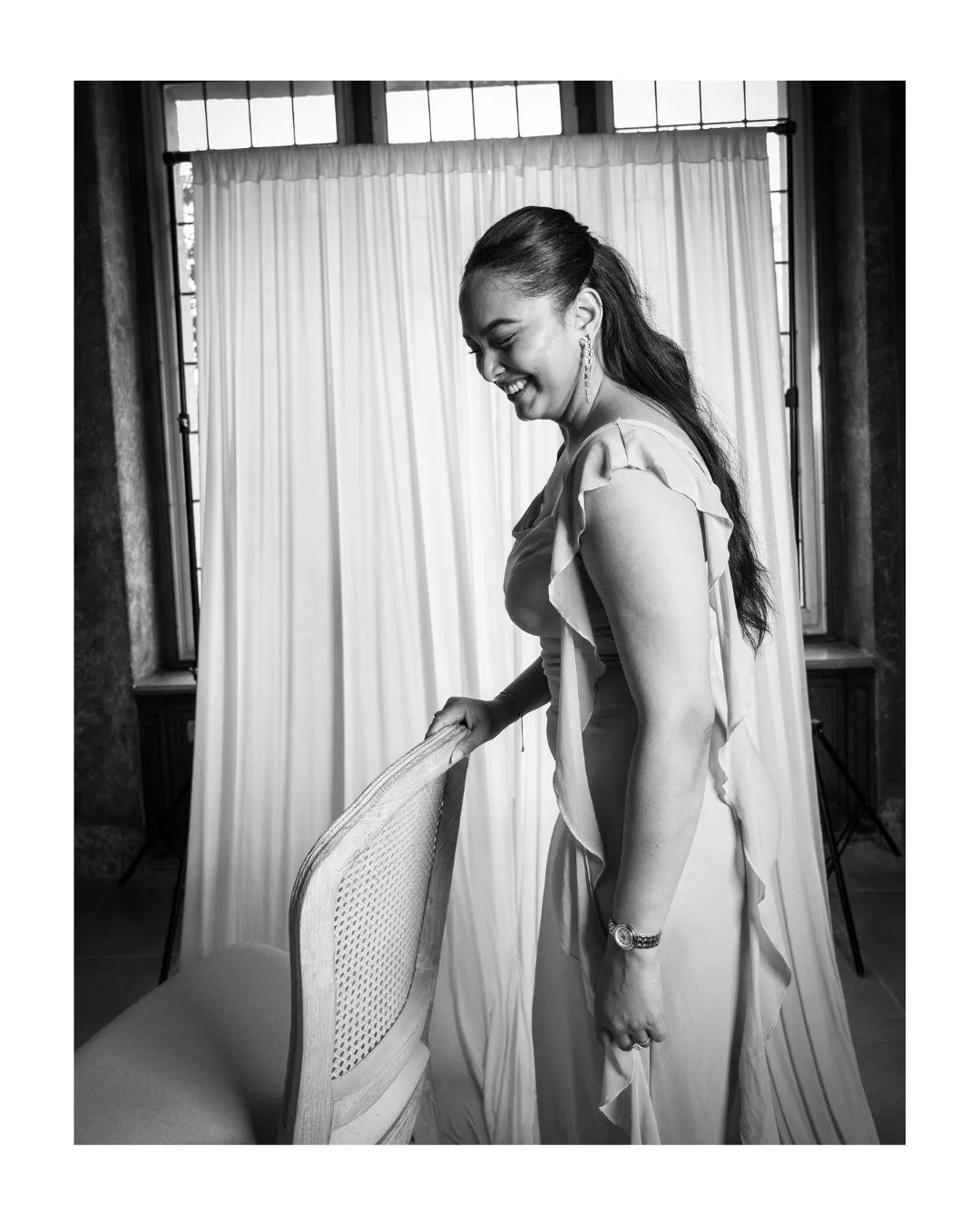 Black and white portrait of a smiling woman holding the back of a chair beside a draped backdrop