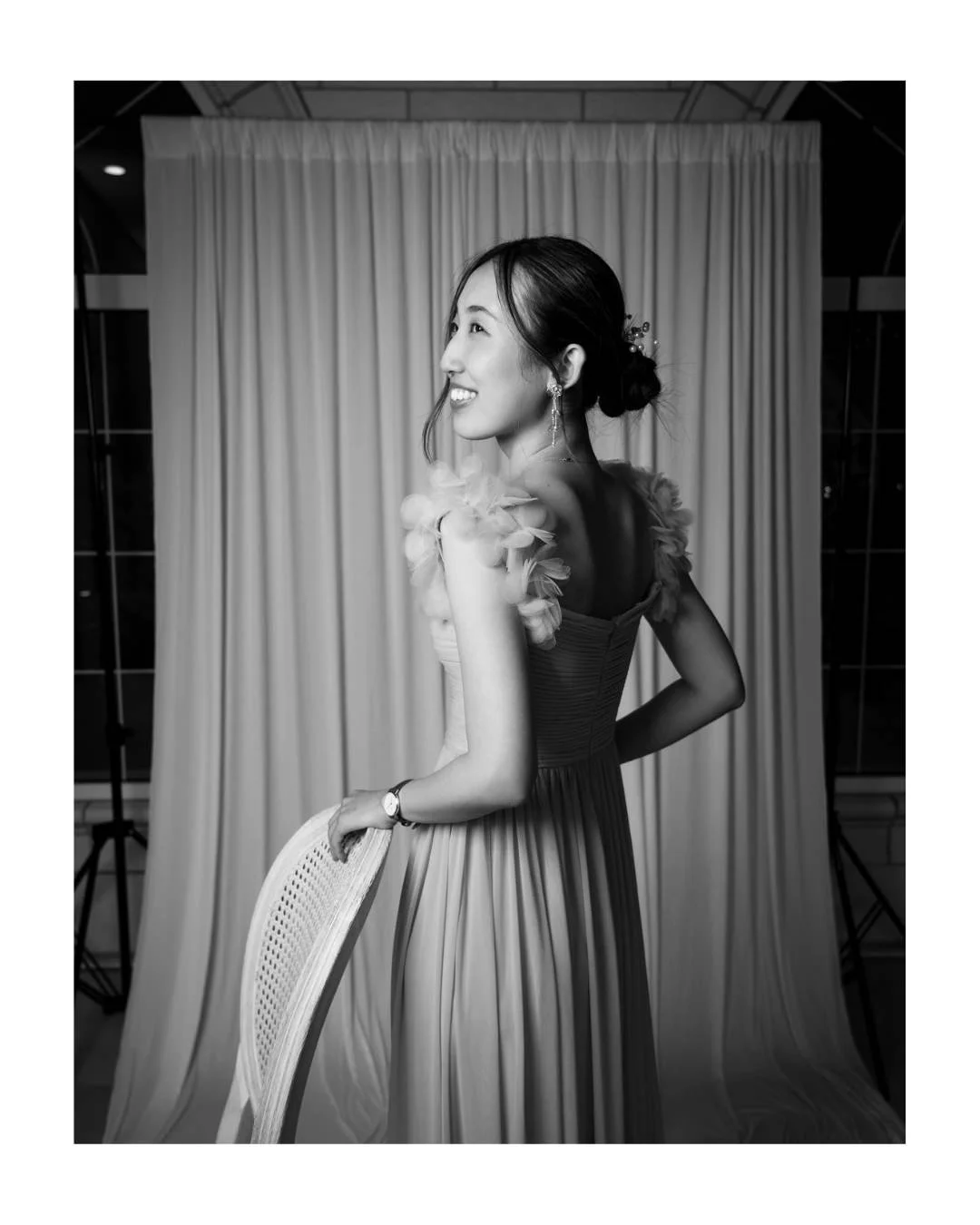 Black and white portrait of a smiling woman in a ruffled-shoulder gown, hand on a cane-back chair, glancing over her shoulder