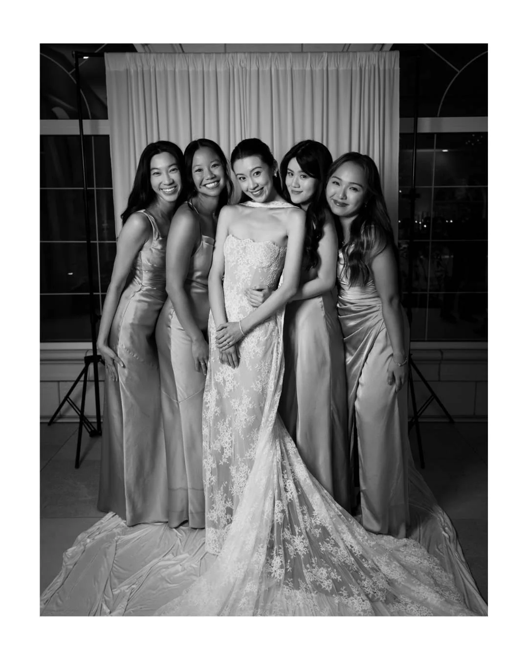Black and white portrait of five women in a bridal party wearing elegant gowns standing together with a bride in front of a draped backdrop